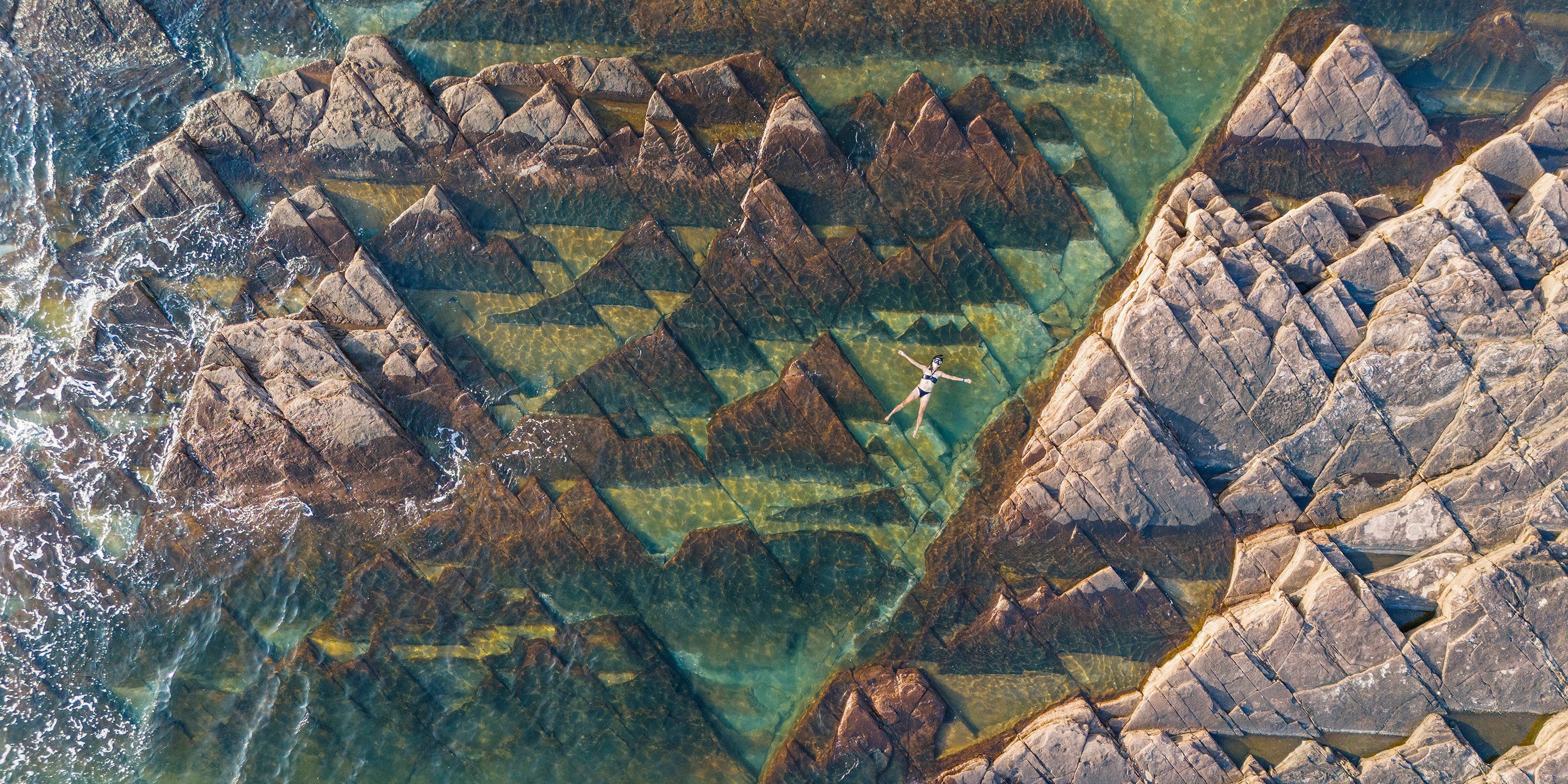 An aerial view of a person floating on their back in shallow water, among triangle-shaped rock formations.