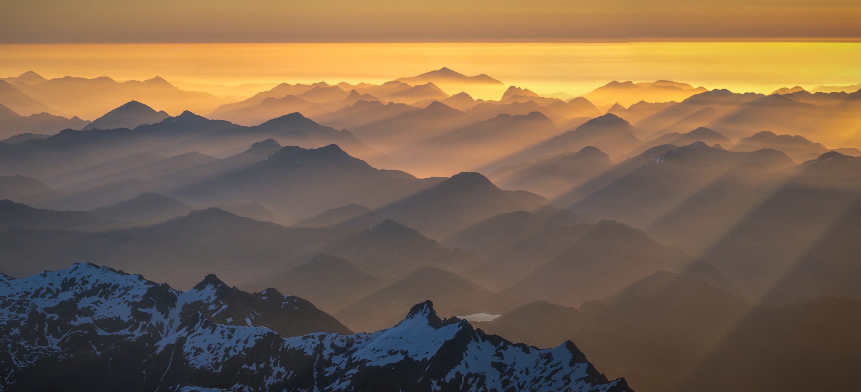 Rays of sunlight filter through fog and valleys among many mountain peaks, seen from above.