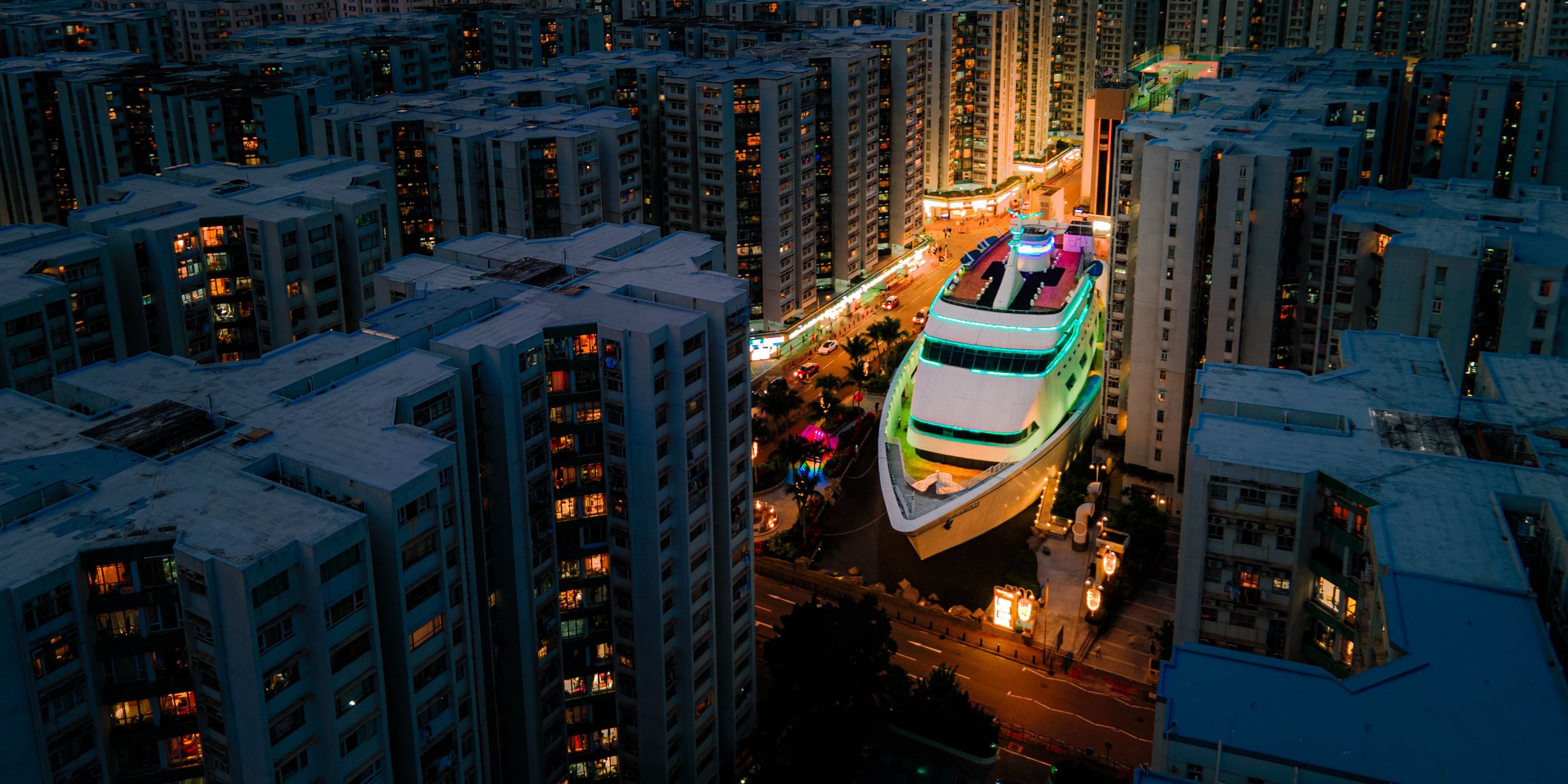An elevated view of a large boat-shaped building sitting in a plaza among many other multi-story residential buildings.