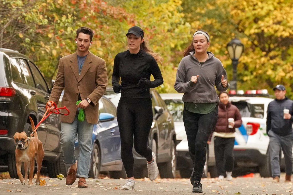 A man walking a tan dog on a leash and two women jogging with a street and trees in the background.