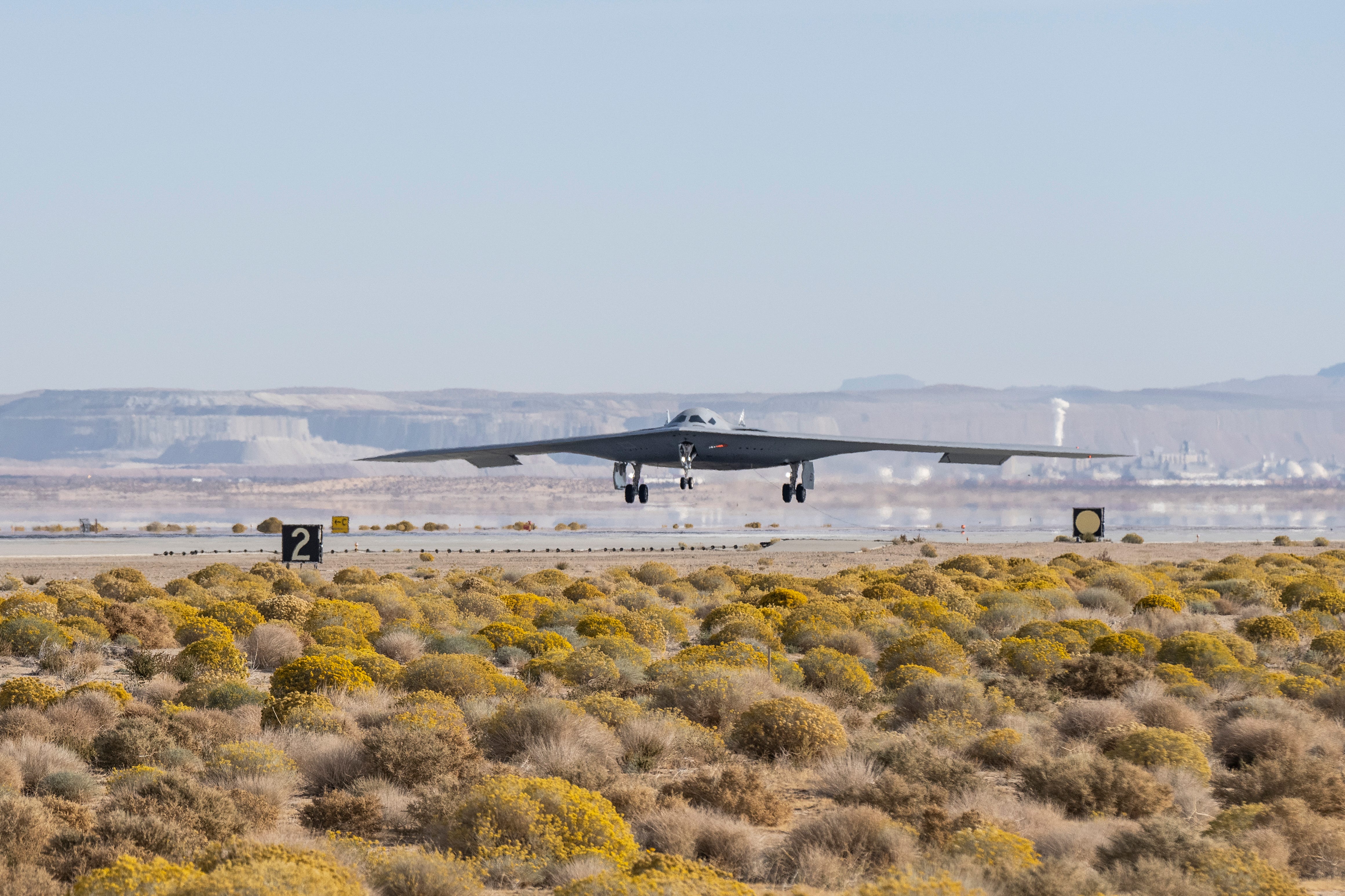 A US Air Force B-21 bomber flies above the tarmac with a blue sky in the background green and yellow desert plans in the foreground.