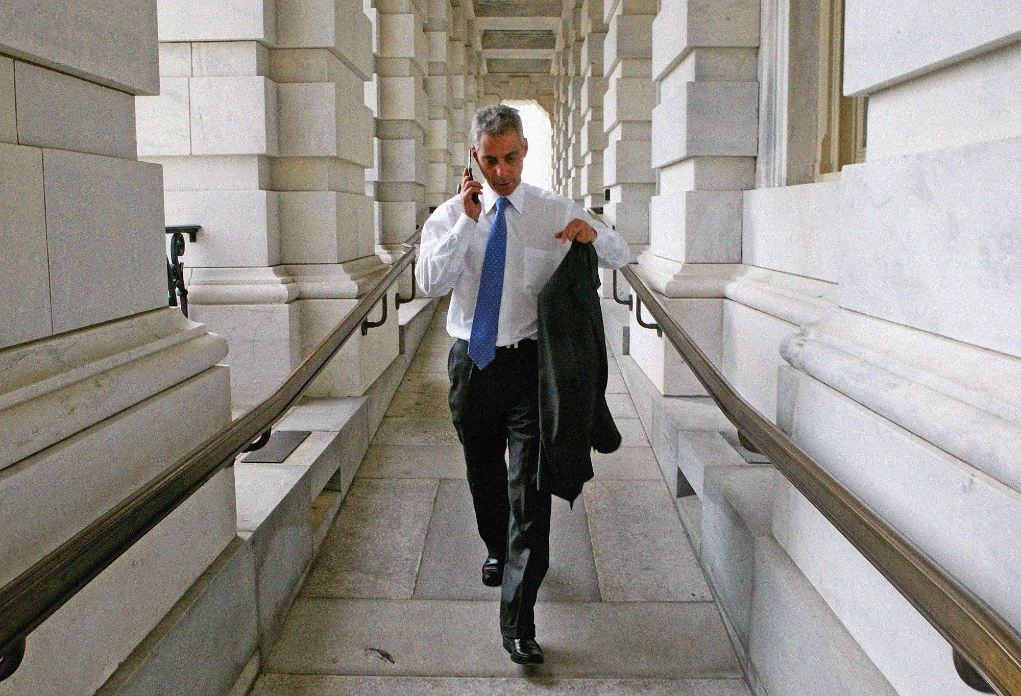 Rahm Emanuel holding a cell phone and his jacket walking in a corridor outside of the U.S. Capitol.