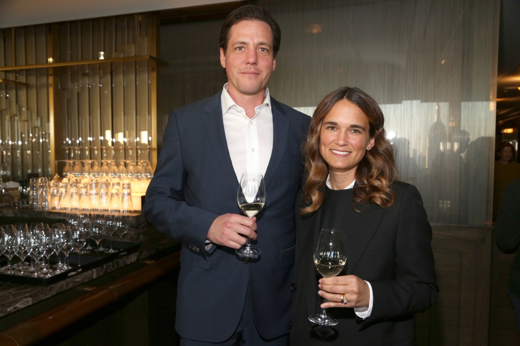 A man in a suit and a woman in a black jacket smiling and holding glasses of white wine.