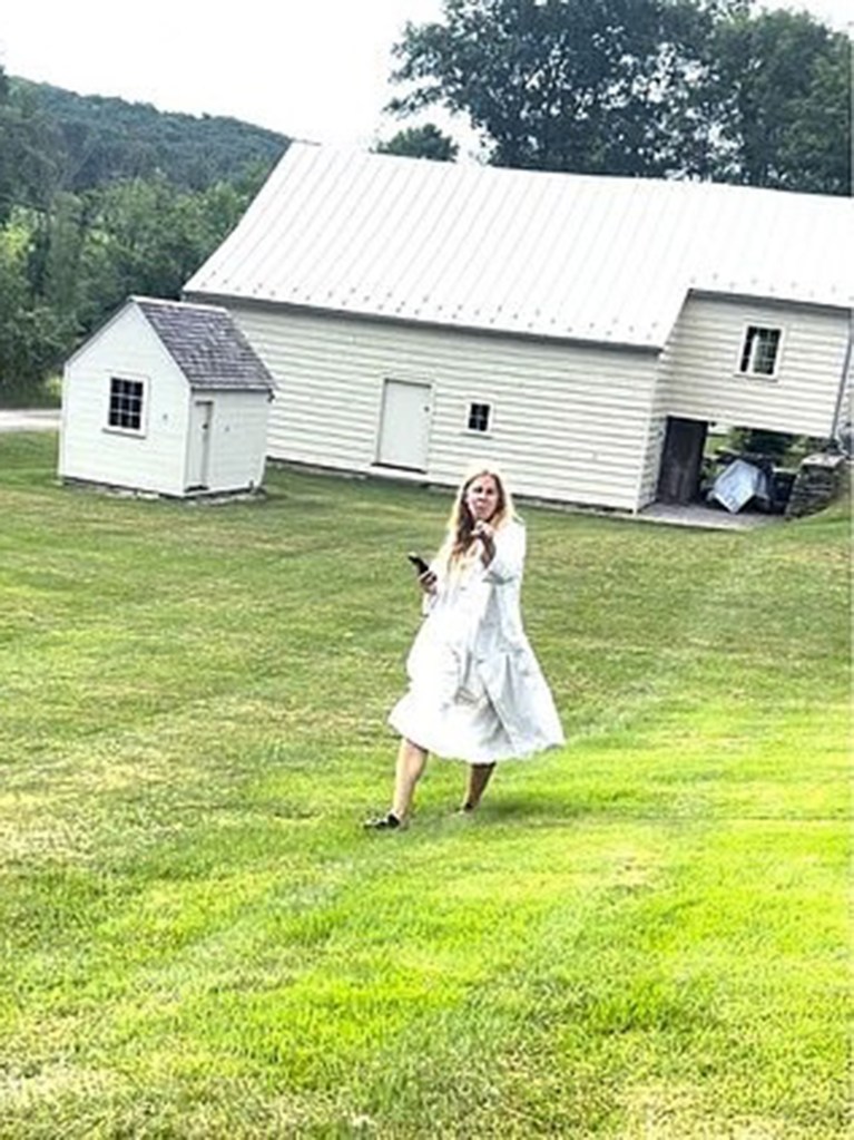 Woman in a white dress and black shoes stands in a grassy field, holding a phone, with a white house and shed behind her.