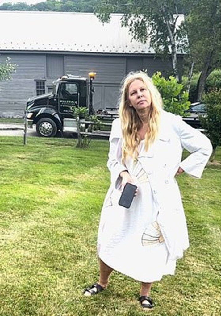 A woman with long blonde hair and a white coat holds a phone, standing in a grassy area with a tow truck and barn in the background.