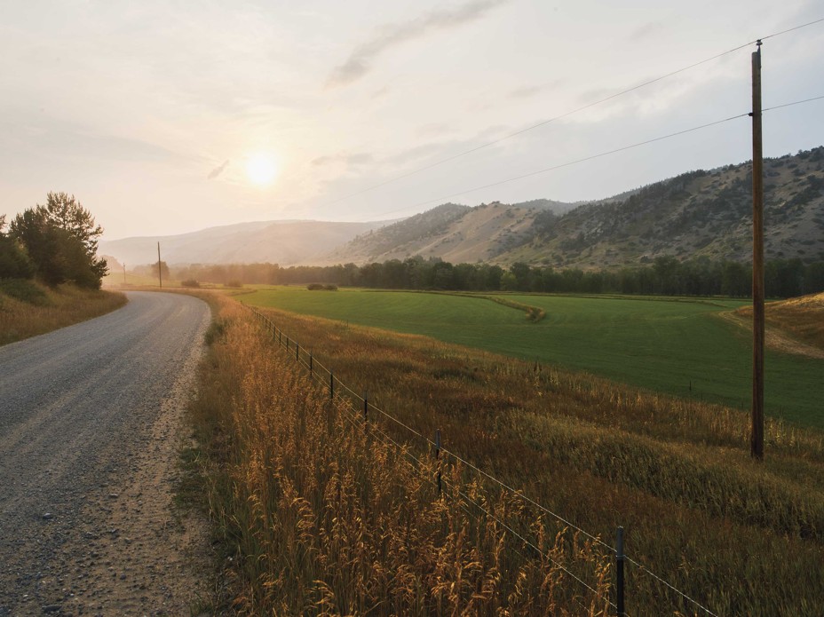 photo of rural road at left along a field bordered on right by green hills, with sun low over horizon