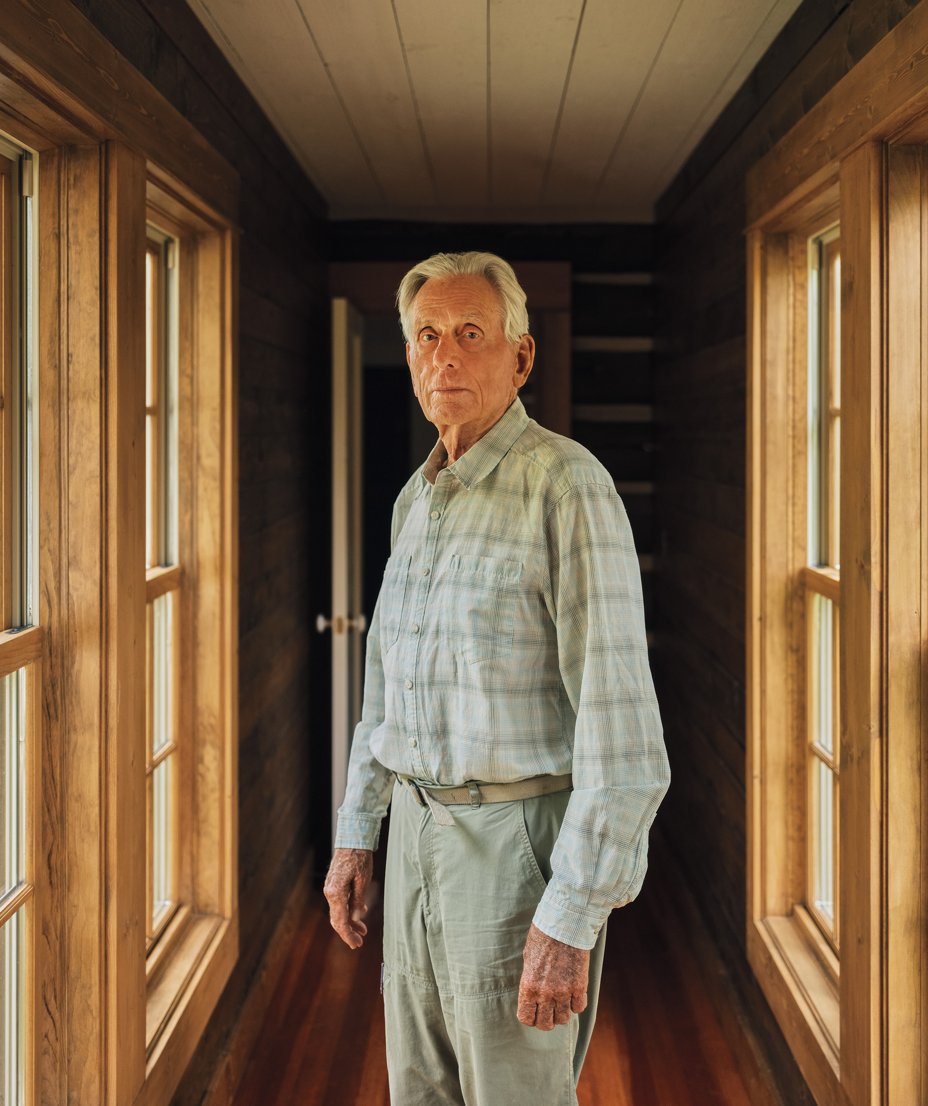 photo of man with white hair wearing plaid shirt and light-green pants standing in hallway with large windows on both sides