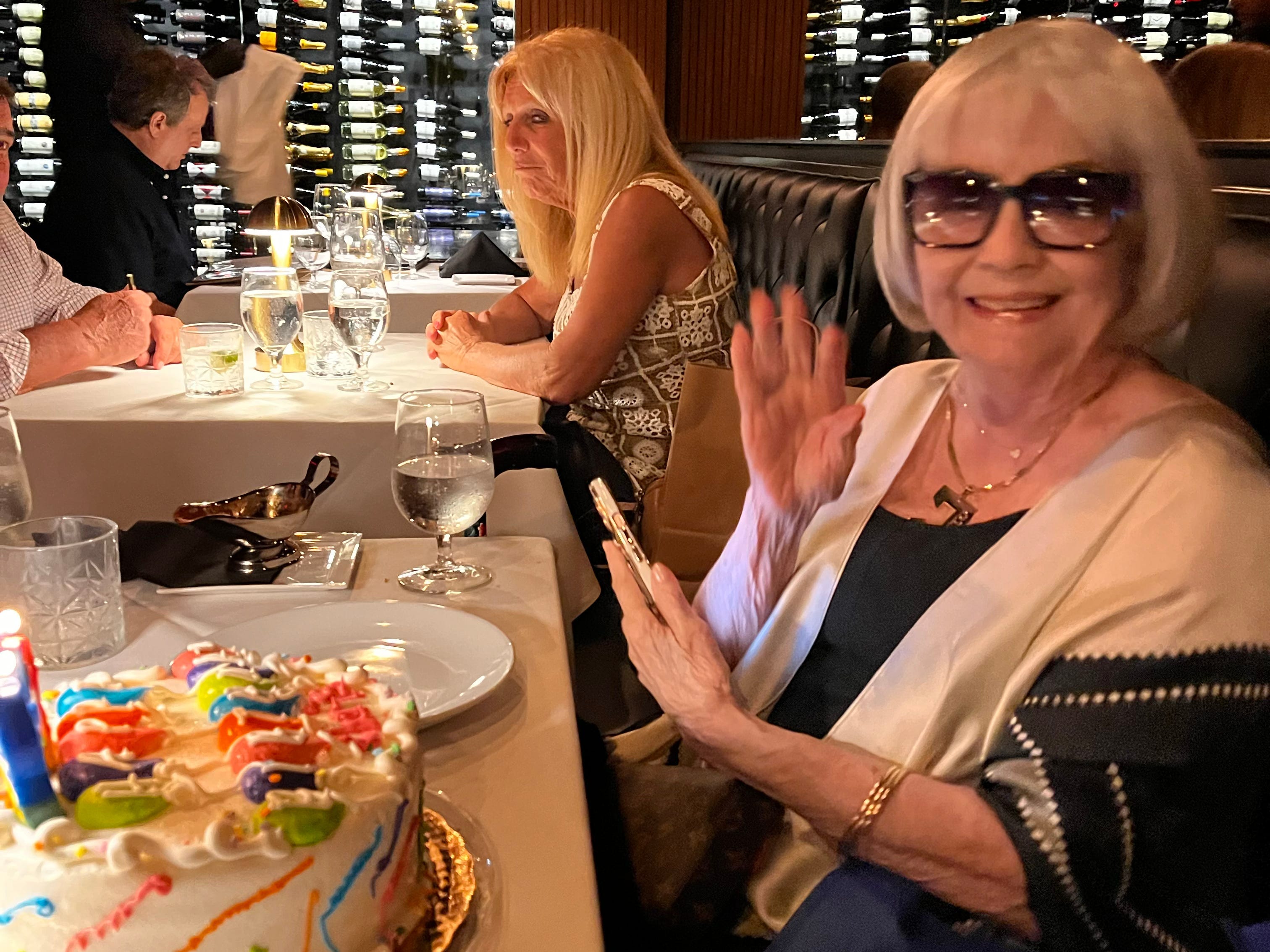An older woman is sitting at a dinner table with a birthday cake.