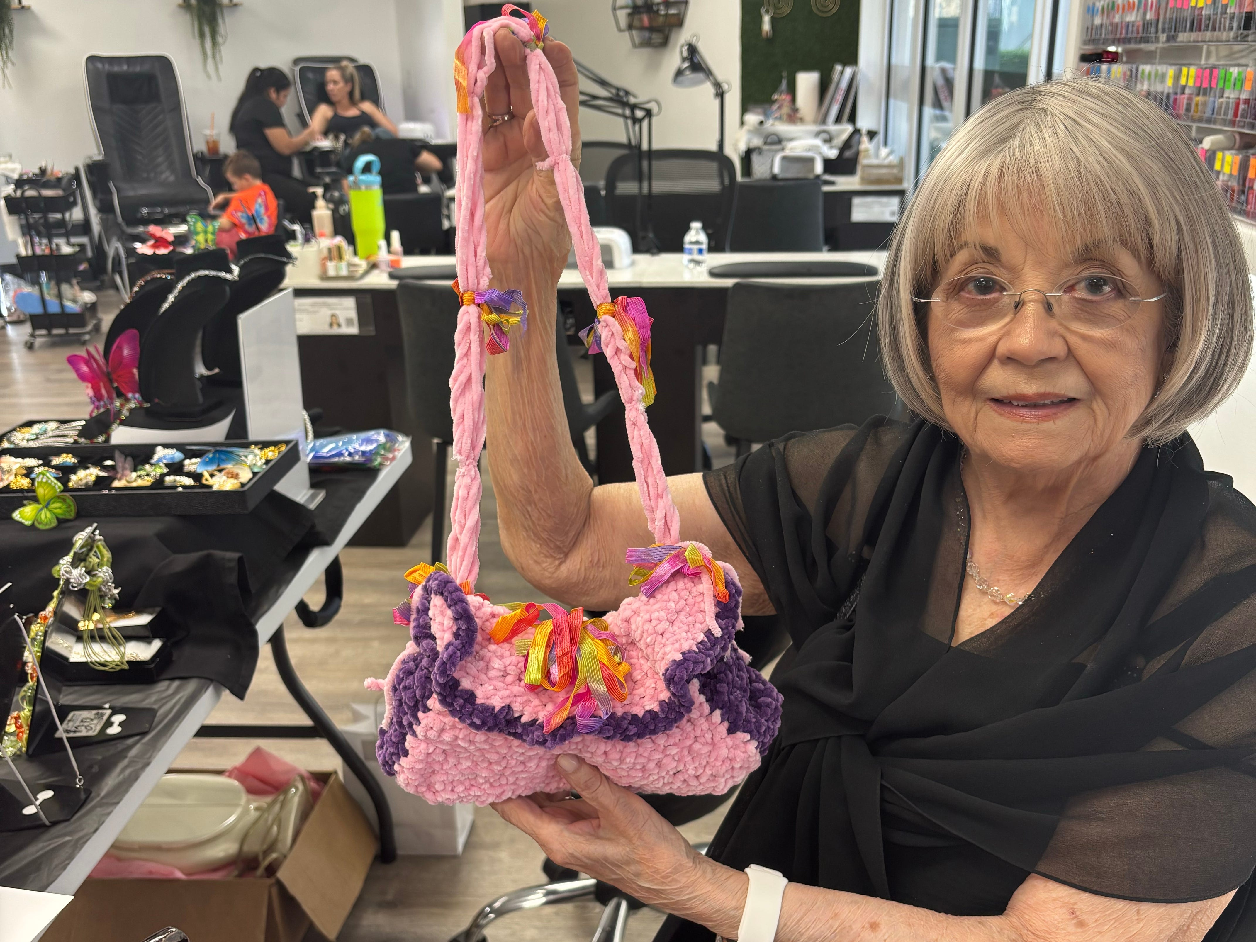 An older woman is holding up a pink, knitted purse she made