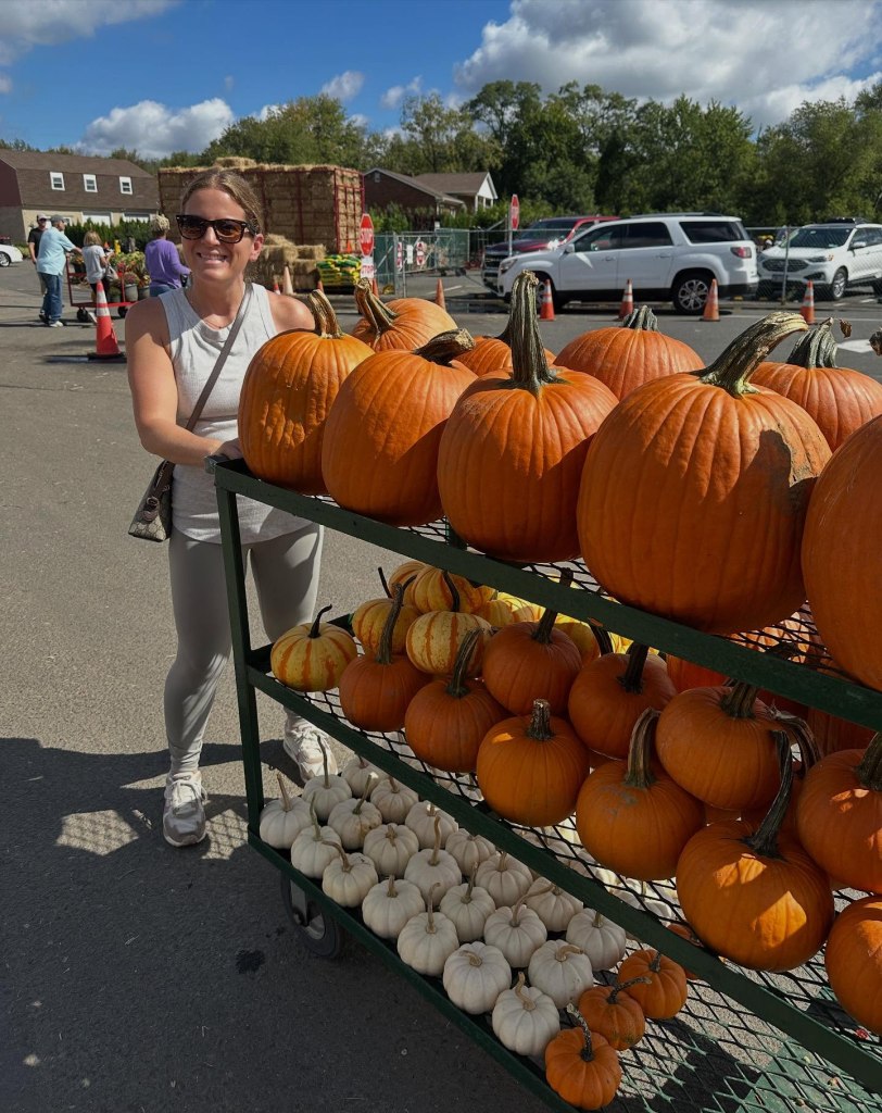 Emily Gonzalez, co-owner of The Entry Edit, poses next to a cart of pumpkins, including large orange ones, small striped ones, and white ones.