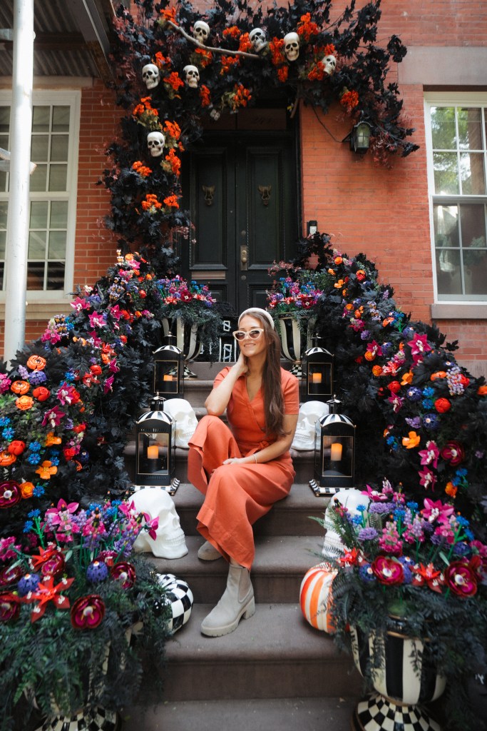 A woman sits on the stoop of a building decorated for Halloween with black and orange foliage, white skulls, and colorful flowers.