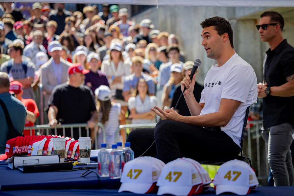 Charlie Kirk speaking into a microphone at an outdoor event with a crowd in the background.