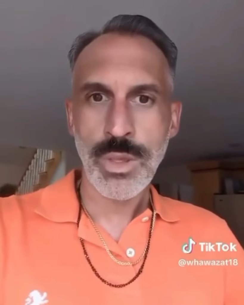 A man with dark hair, a mustache, and a gray beard is wearing an orange shirt and gold chain necklaces.