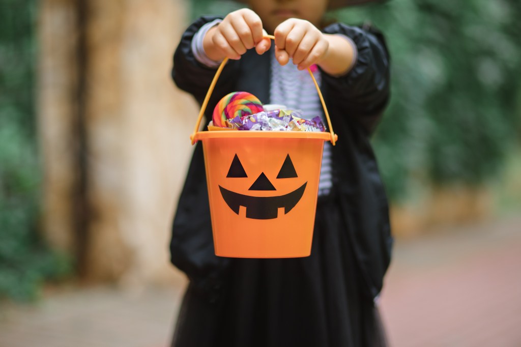 Little cute girl in witch costume holding jack-o-lantern pumpkin bucket with candies and sweets.