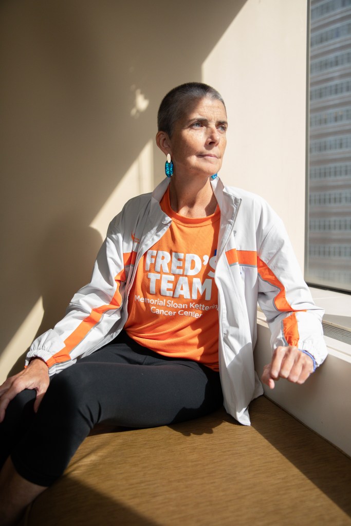 Liz Healy, a woman with short gray hair, in an orange t-shirt and white jacket, sits looking out a window.