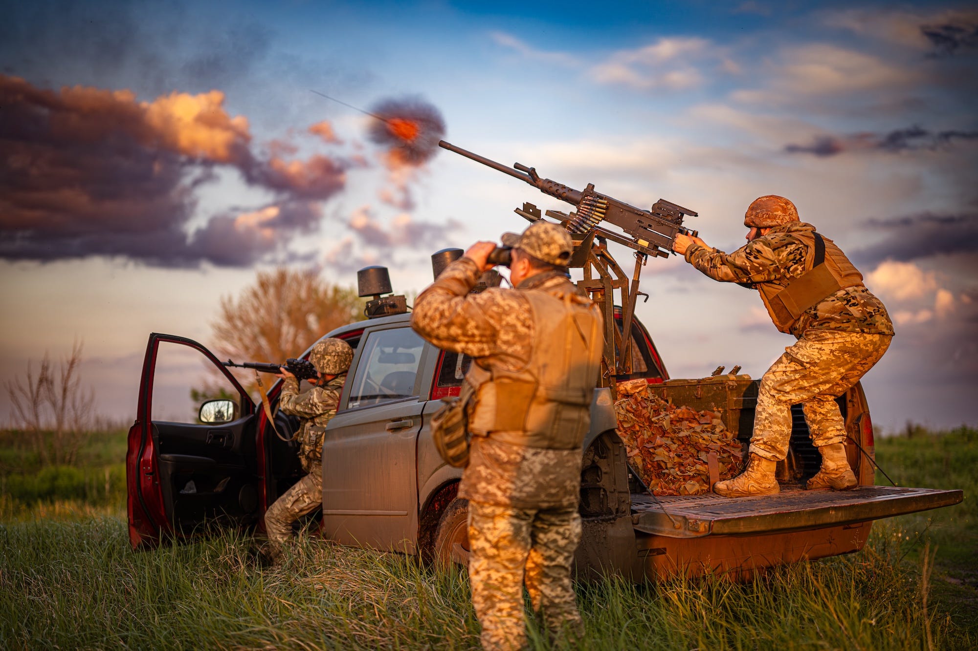 A man on the back of a truck shoots a gun into the air, with another man in the front of the truck and another standing beside it, all wearing camouflage