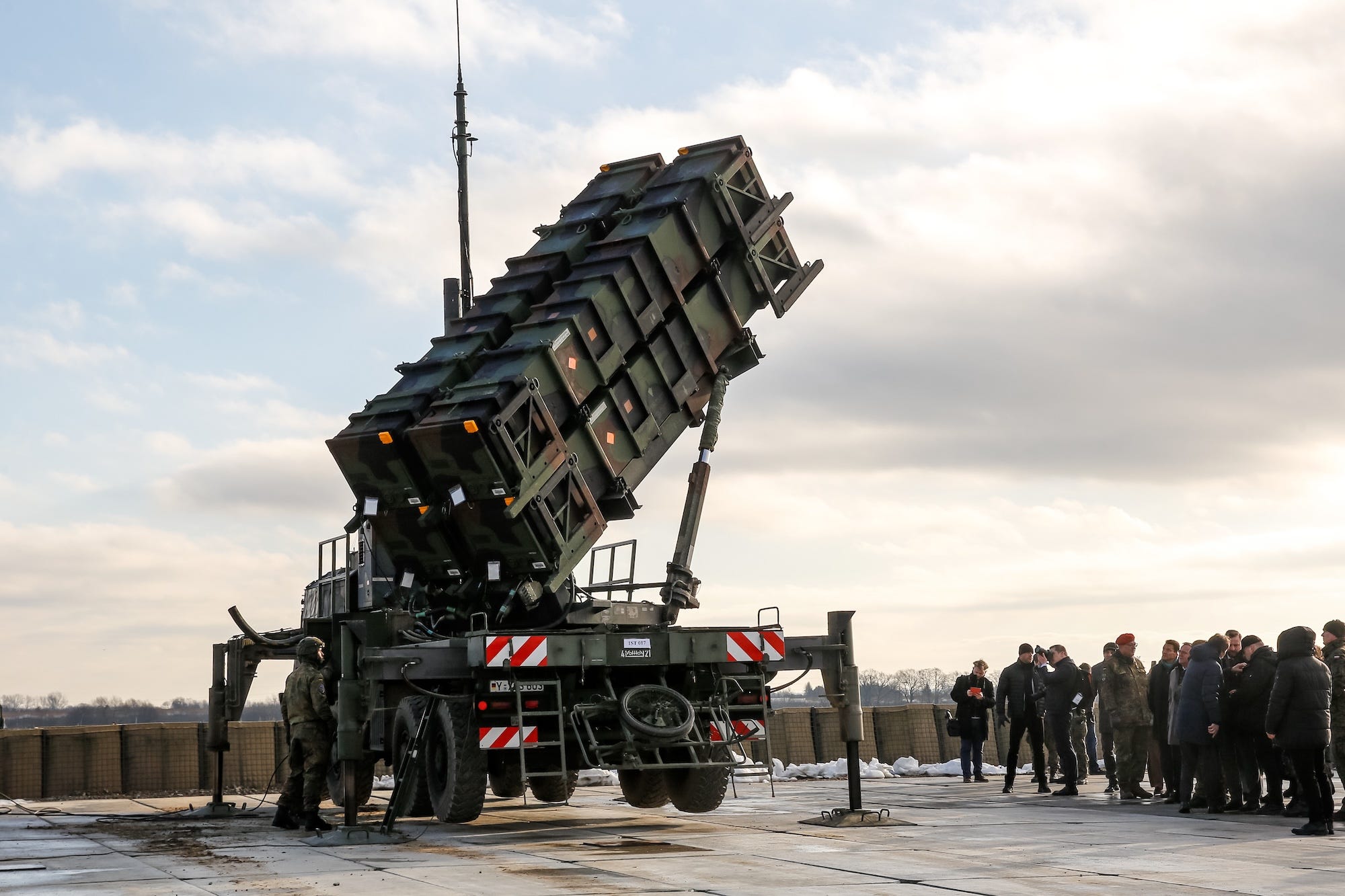 A Patriot air defense system on tarmac with a cloudy skiy behind it and people standing beside it