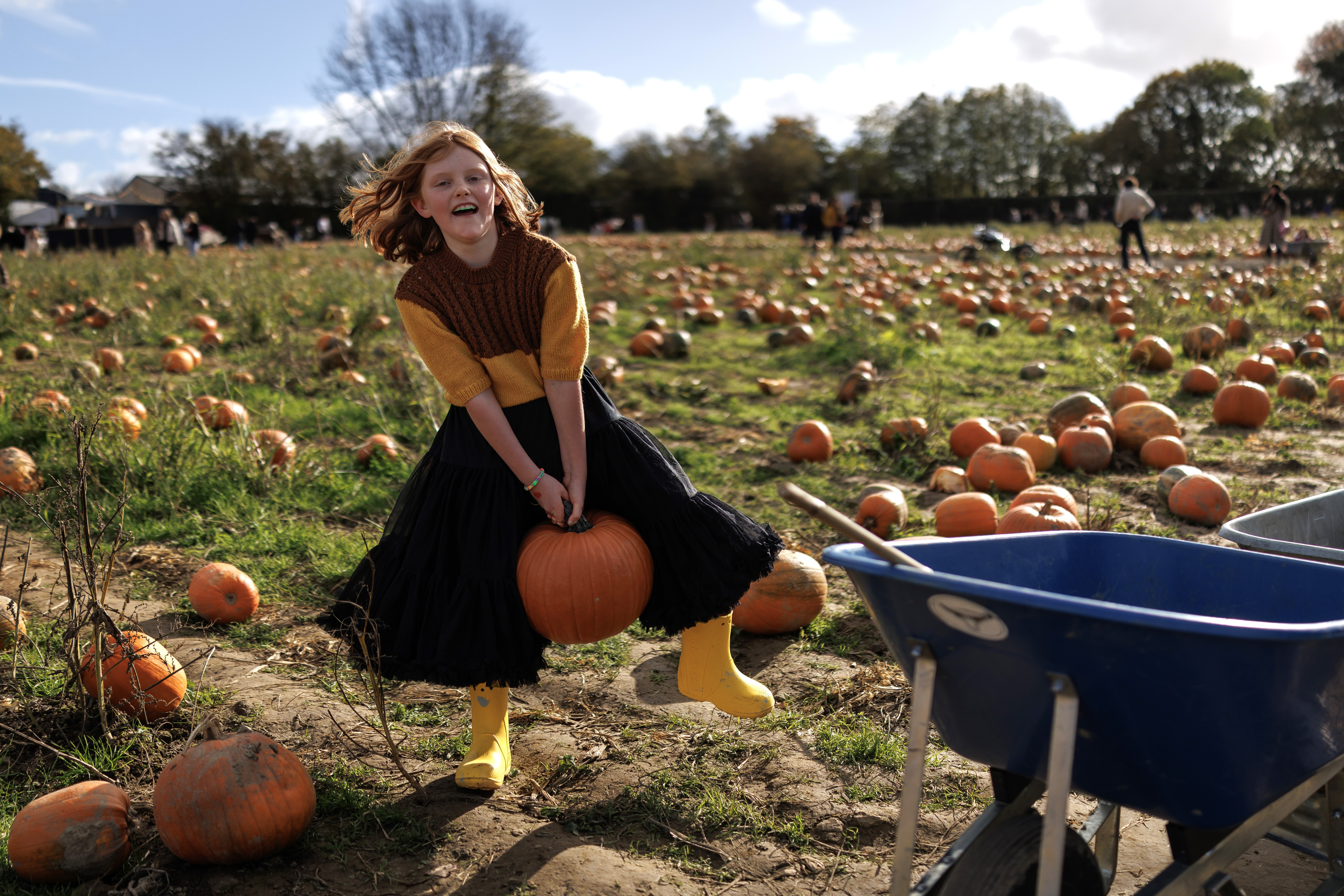 A child smiles while lifting a heavy pumpkin by its stem in a pumpkin patch.