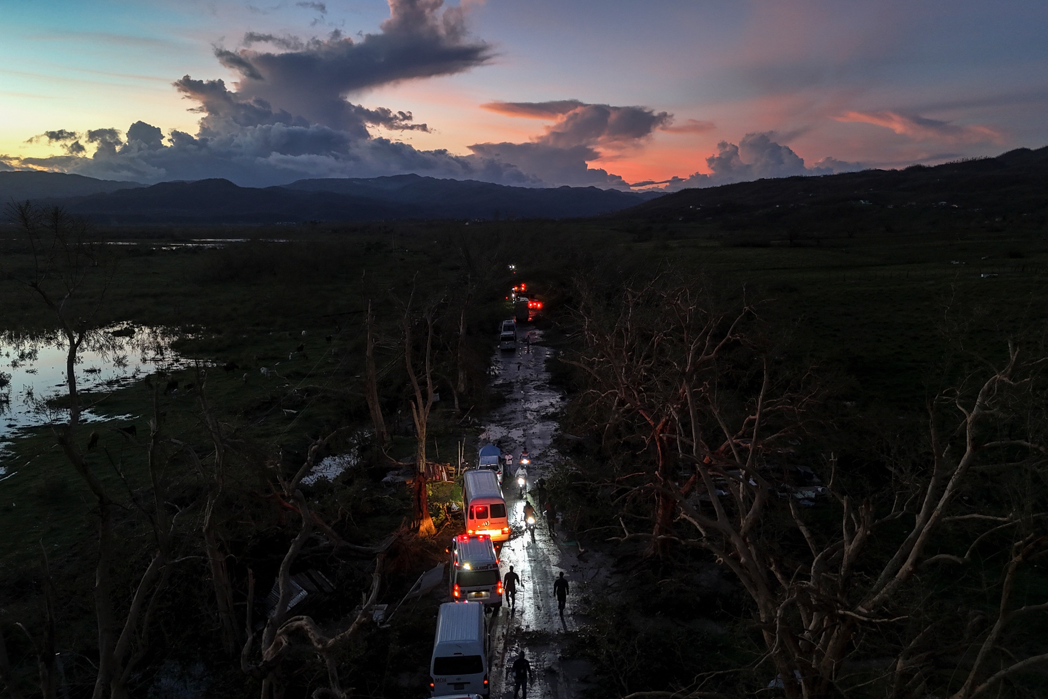 An aid convoy picks its way along a storm-damaged road, past many downed trees.