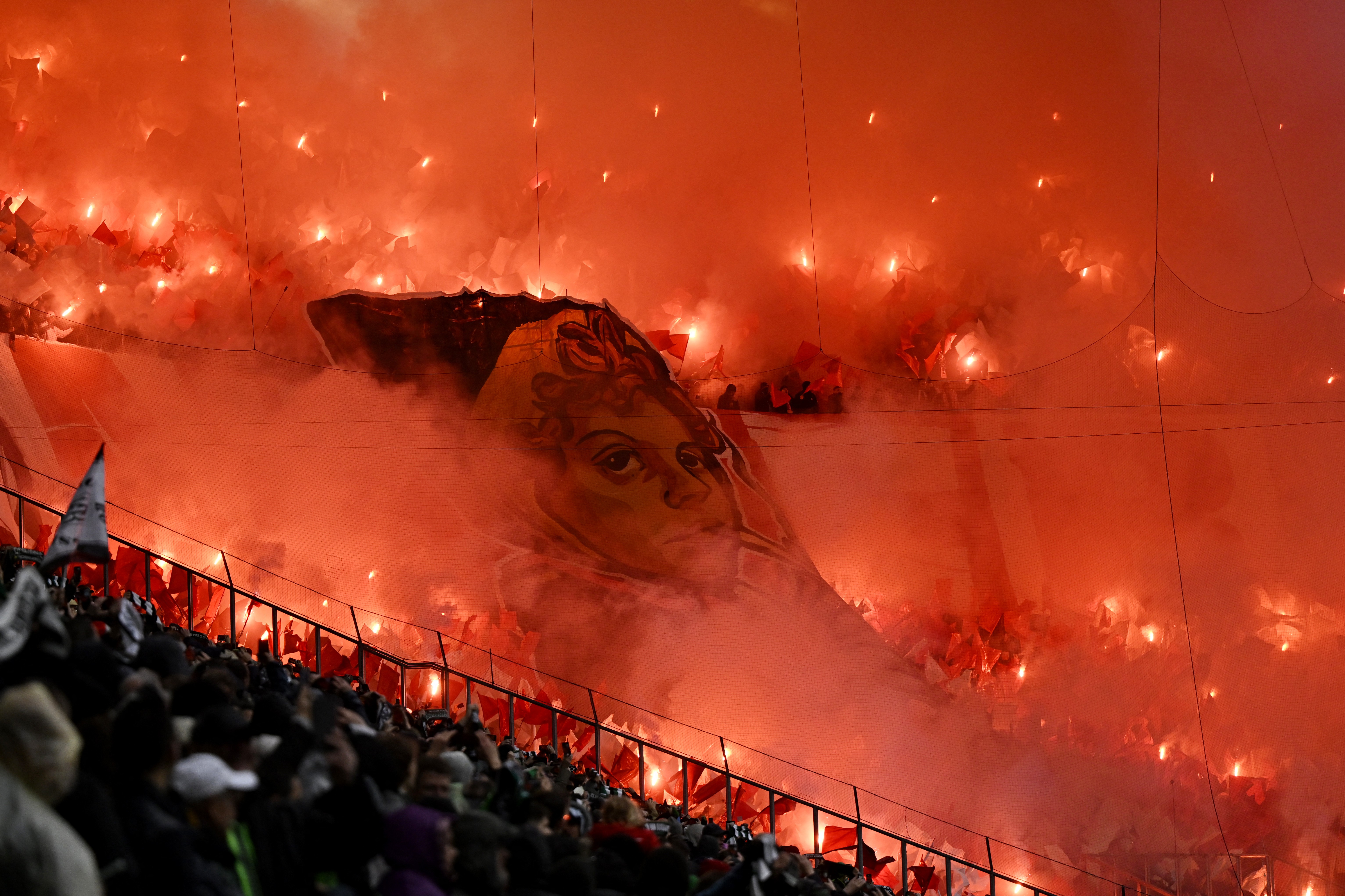 Football fans in a stadium burn flares and display huge banners.