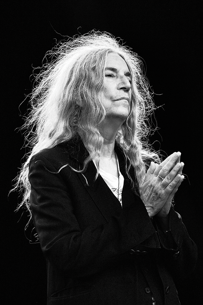 black-and-white photo of woman with long silver hair wearing black jacket and holding hands with palms pressed together in front of her chest