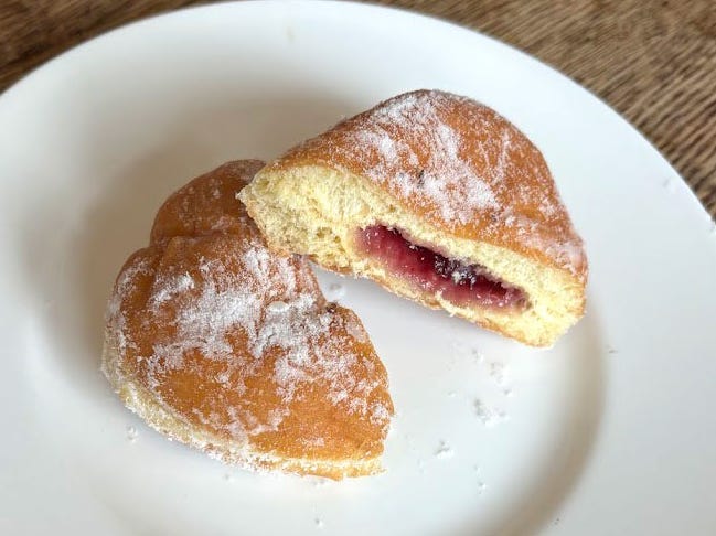 A doughnut with no hole cut in half with red jelly inside. The doughnut is on a white plate.