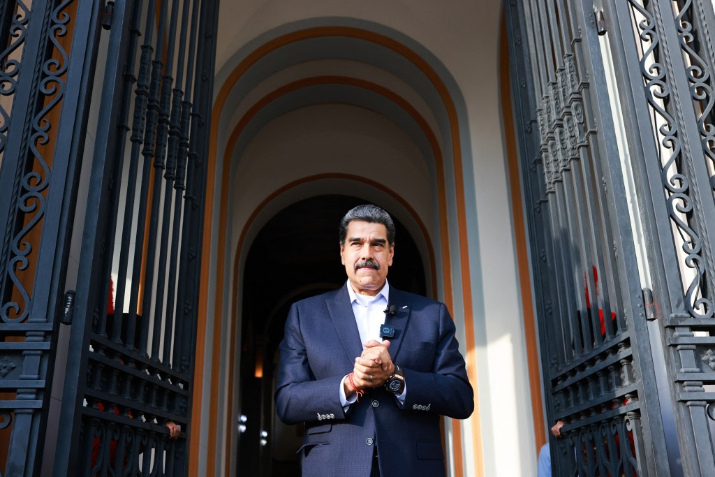 Venezuelan President Nicolas Maduro, wearing a blue suit and white shirt, stands with hands clasped in front of an ornate, open black gate, with arched entrances visible in the background.