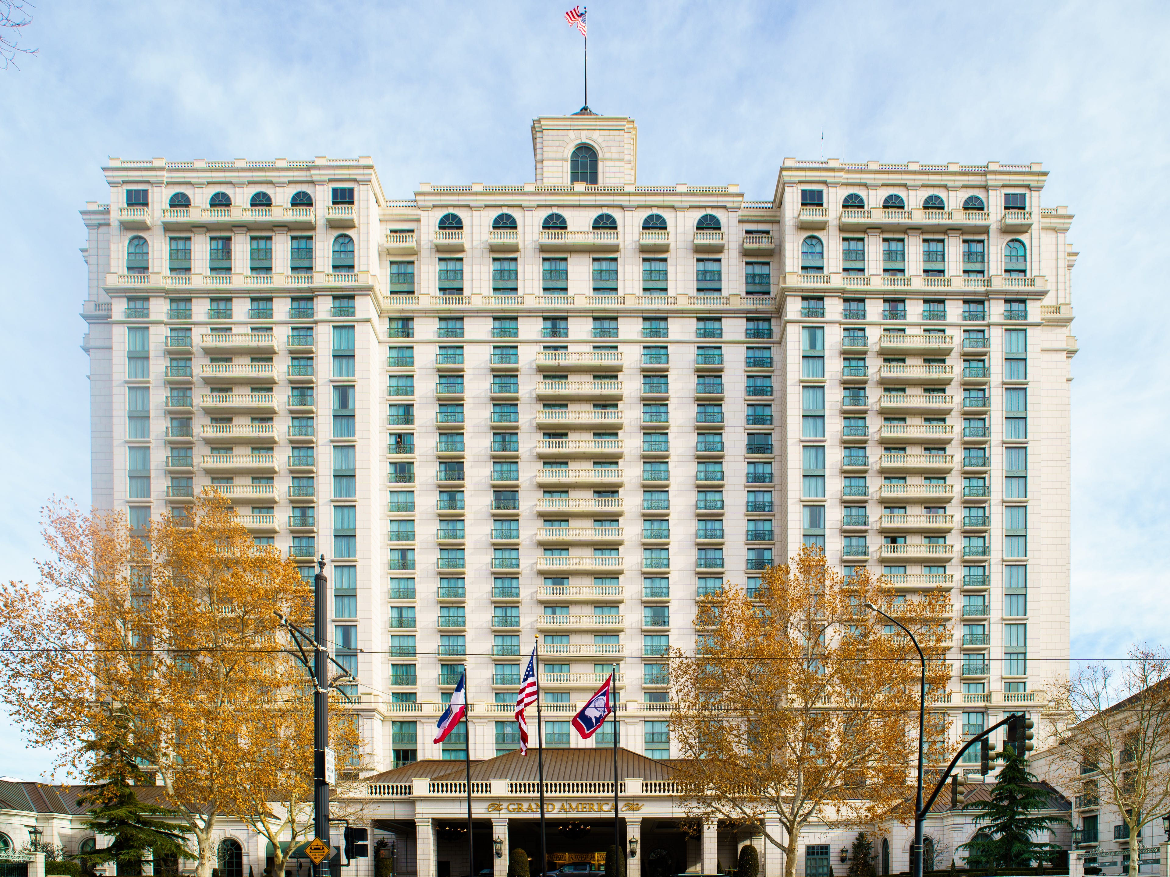 A tall, wide white building, the Grand America Hotel, with flags and trees in the foreground and cloudy skies in the background