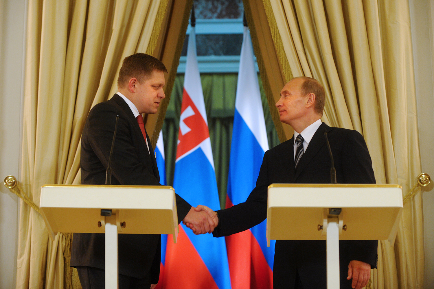 Putin and Fico shake hands. They stand behind identical podiums. Behind them are the flags of Slovakia and Russia framed by curtains.