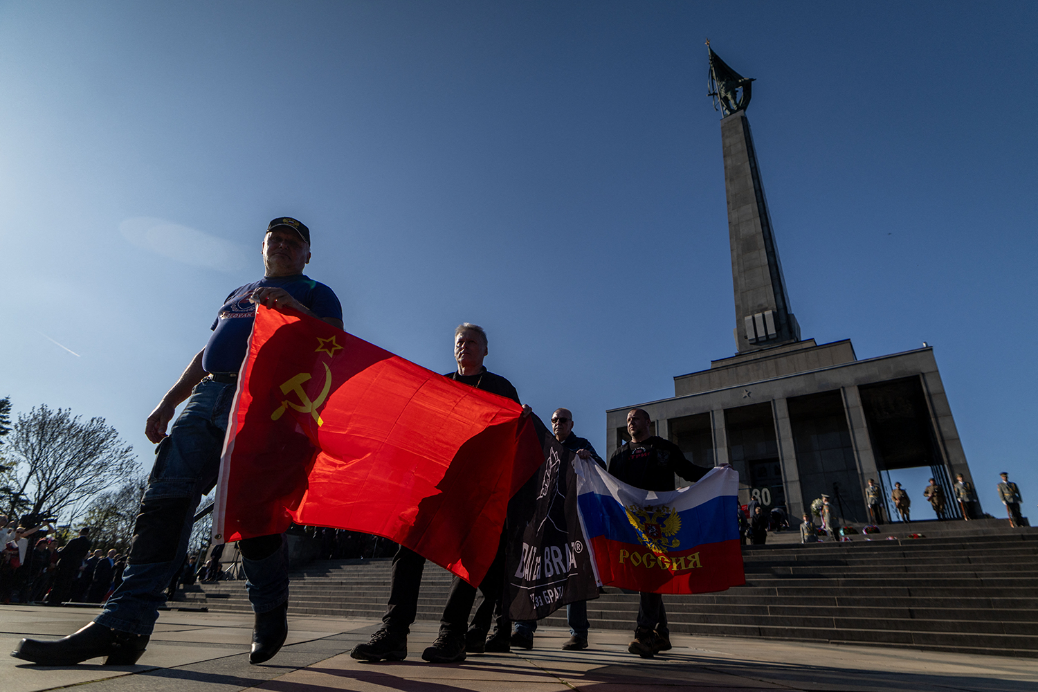 Four men walk through a square, one behind the other, carrying flags. The two men in front carry the flag of the Soviet Union, and the two behind hold the flag of the Russian Federation. An obelisk memorial towers above them in the background.
