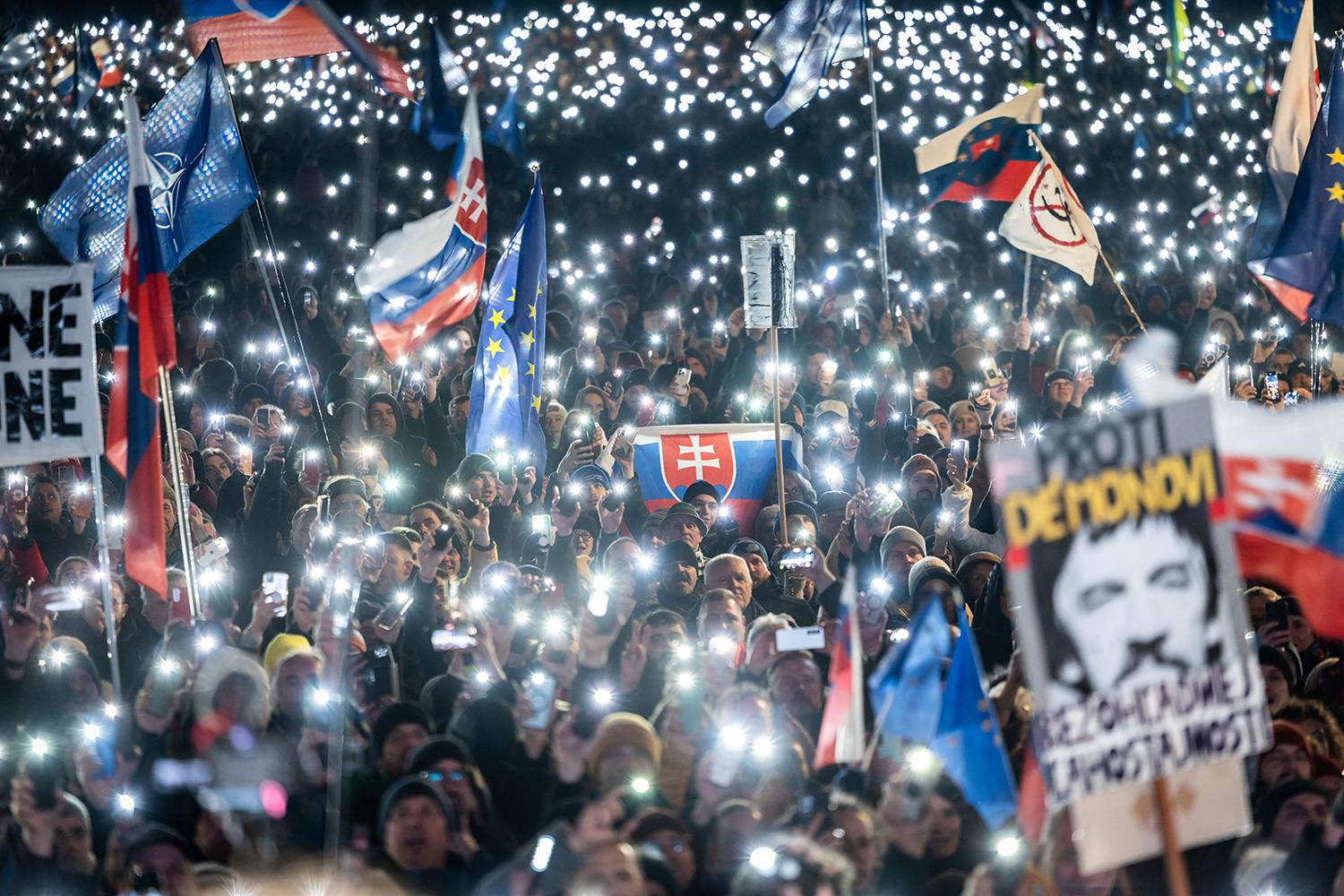 A large crowd of people holds up phone flashlights, as well as Slovak and EU flags and protest posters.