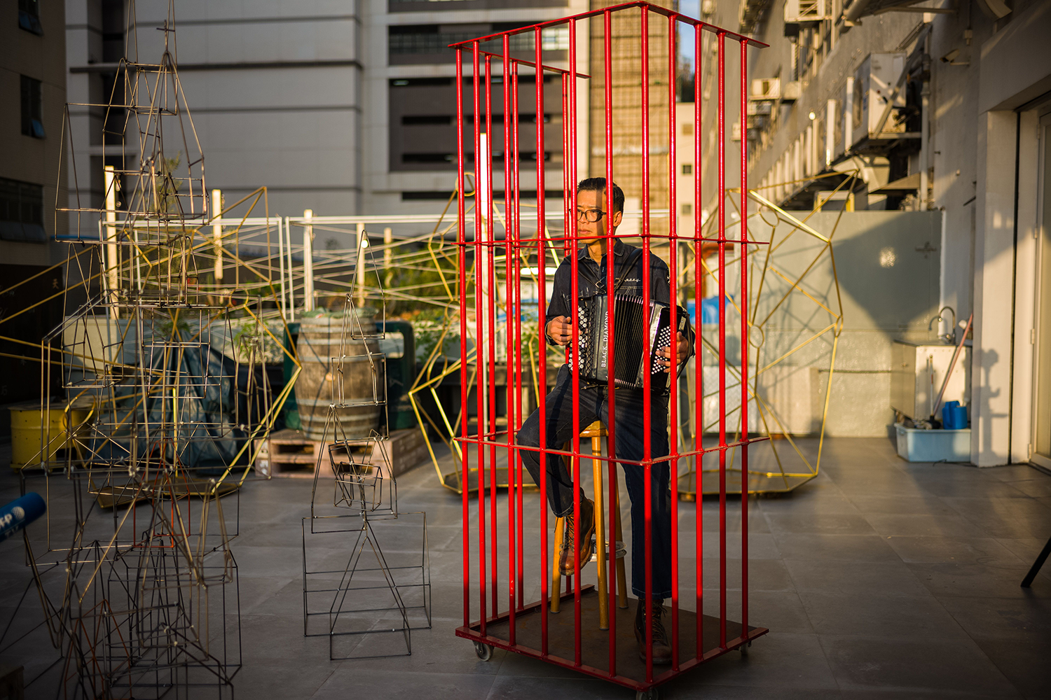 A person sits on a stool and plays an accordion inside a bright red cage. Wire formations and various building supplies are outside of the cage.