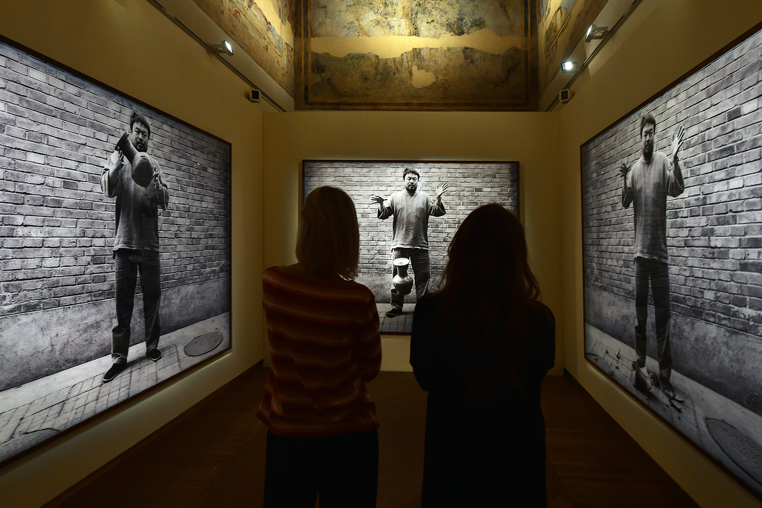 Two people stand before three large images, each on displayed on its own wall. In the first, a man holds an old vase; in the second, the vase hangs in midair as he drops it; and in the third, the shattered pieces lie at his feet.