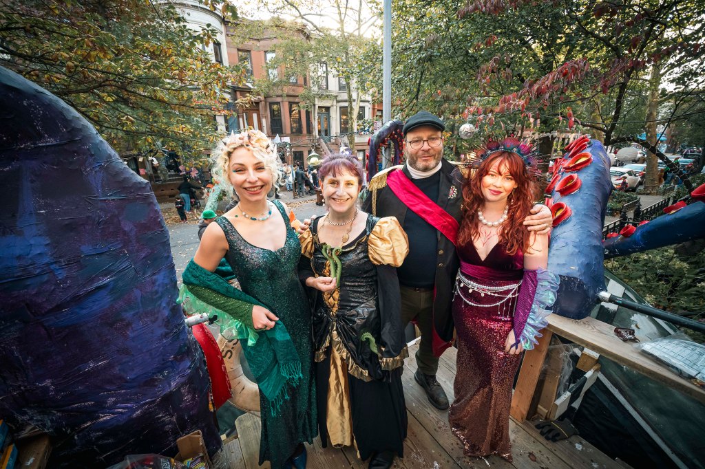 Four adults in Halloween costumes pose in front of a Brooklyn brownstone, with decorations visible in the background.