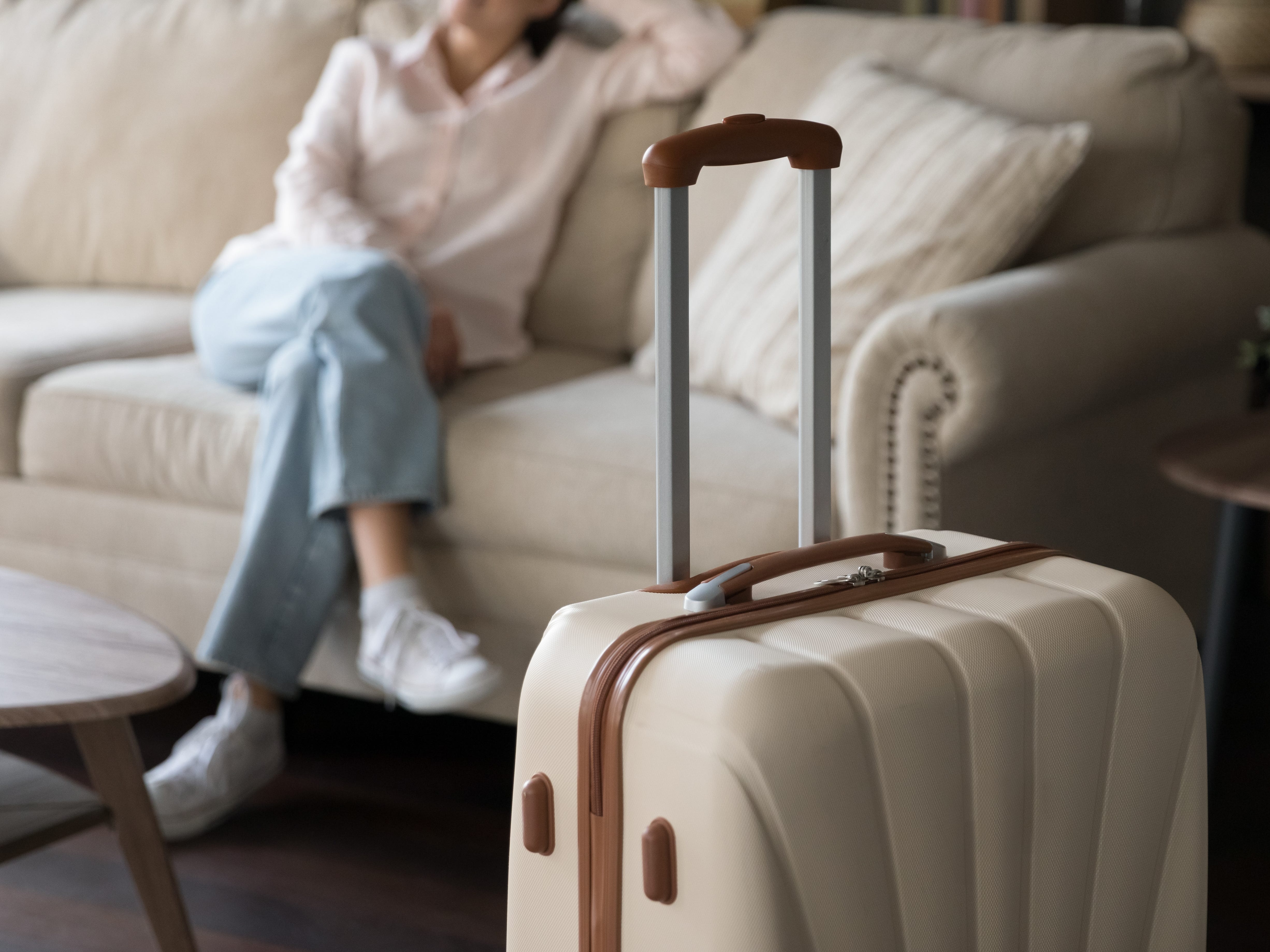 A beige and brown suitcase in a living room, with a woman sitting cross-legged on the couch in the background.