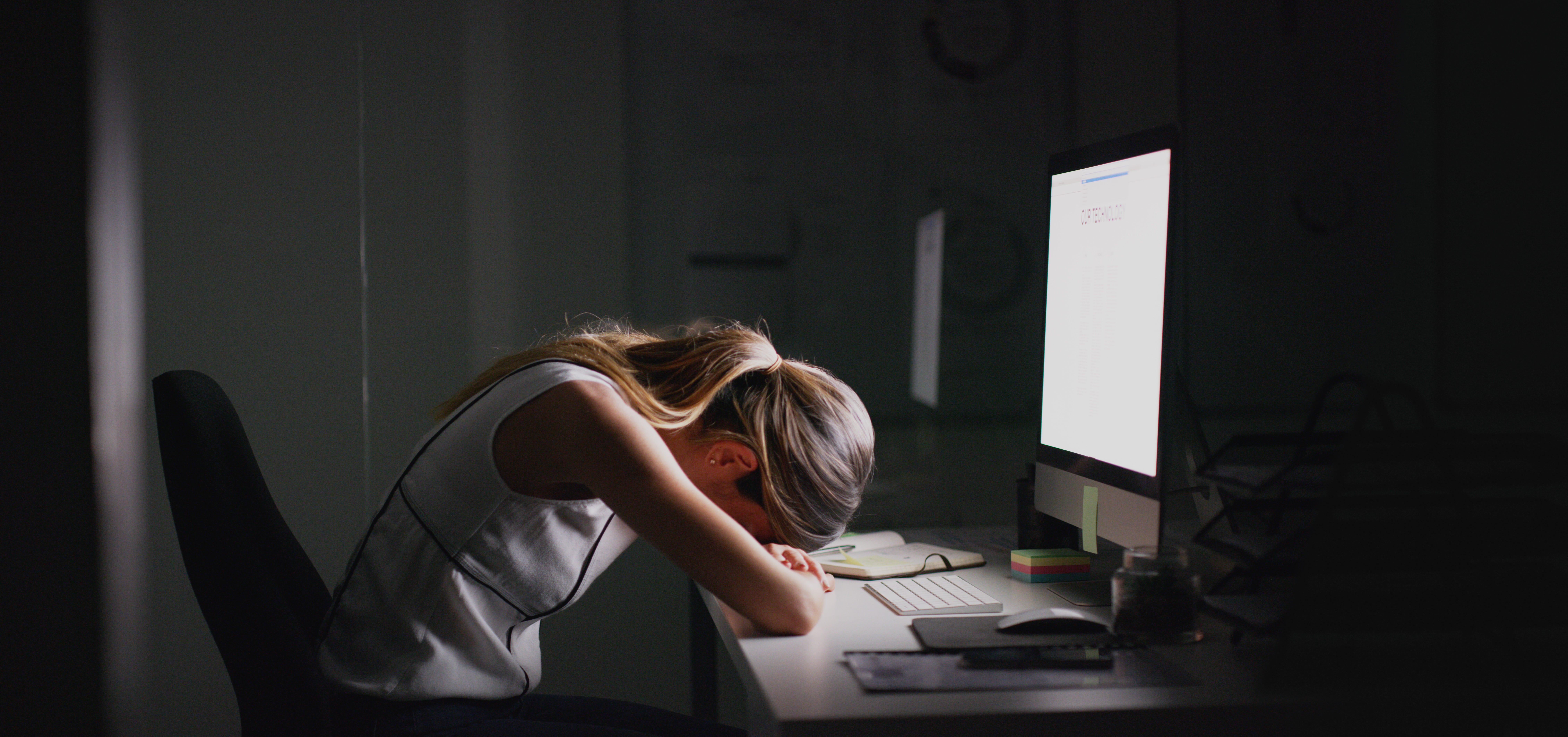 A woman lit by her computer puts her head down on her desk
