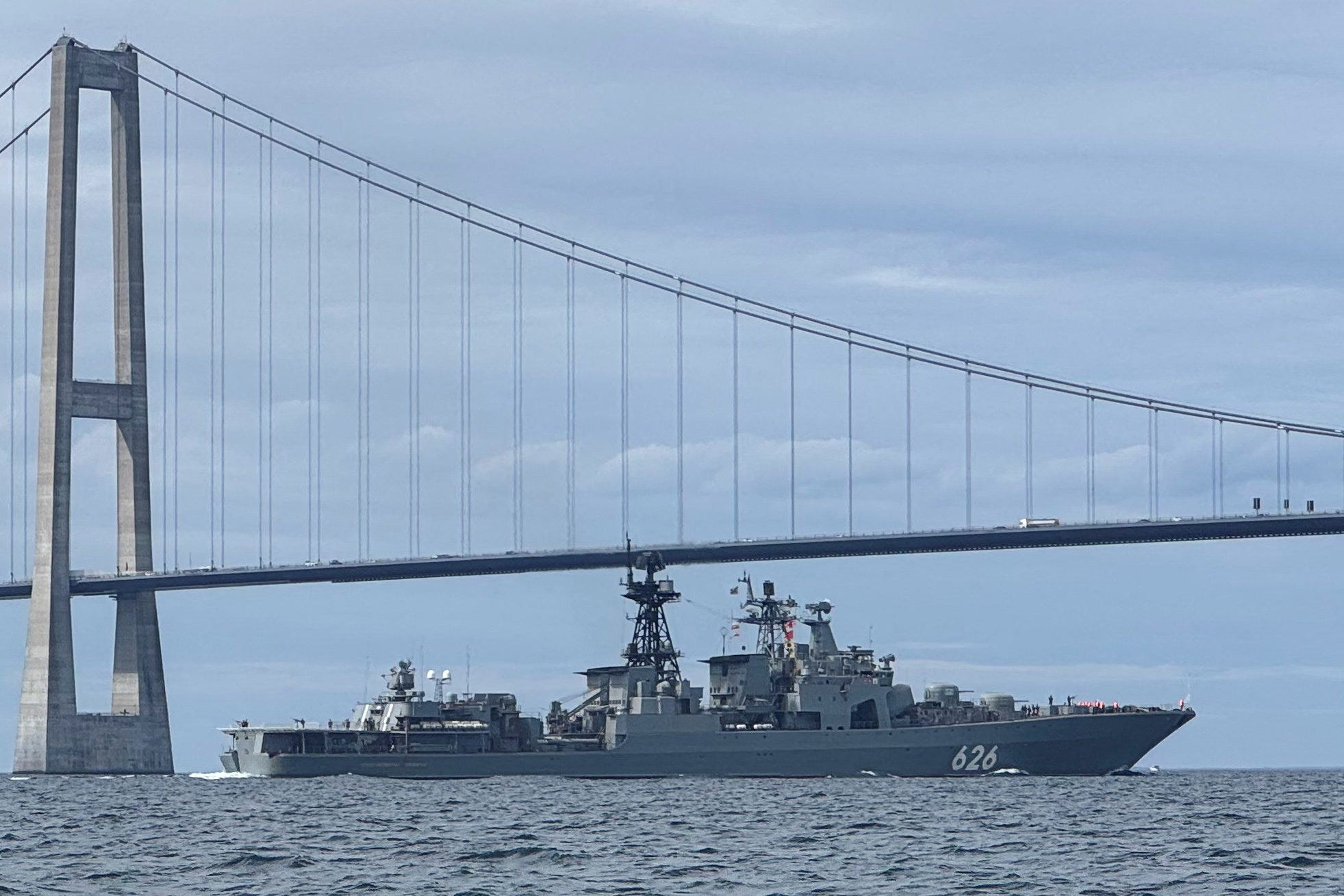 A grey navy vessel in the sea in front of a large grey bridge