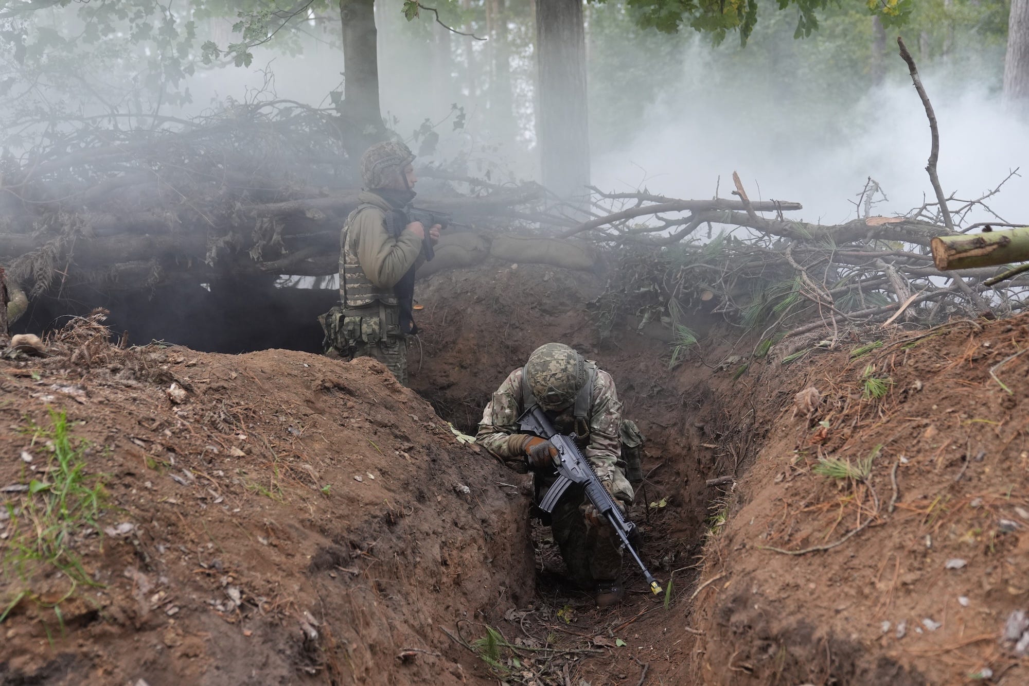 A figure in camouflage gear kneels in a trench while holding a firearm, with smoky air and piled-up branches behind him
