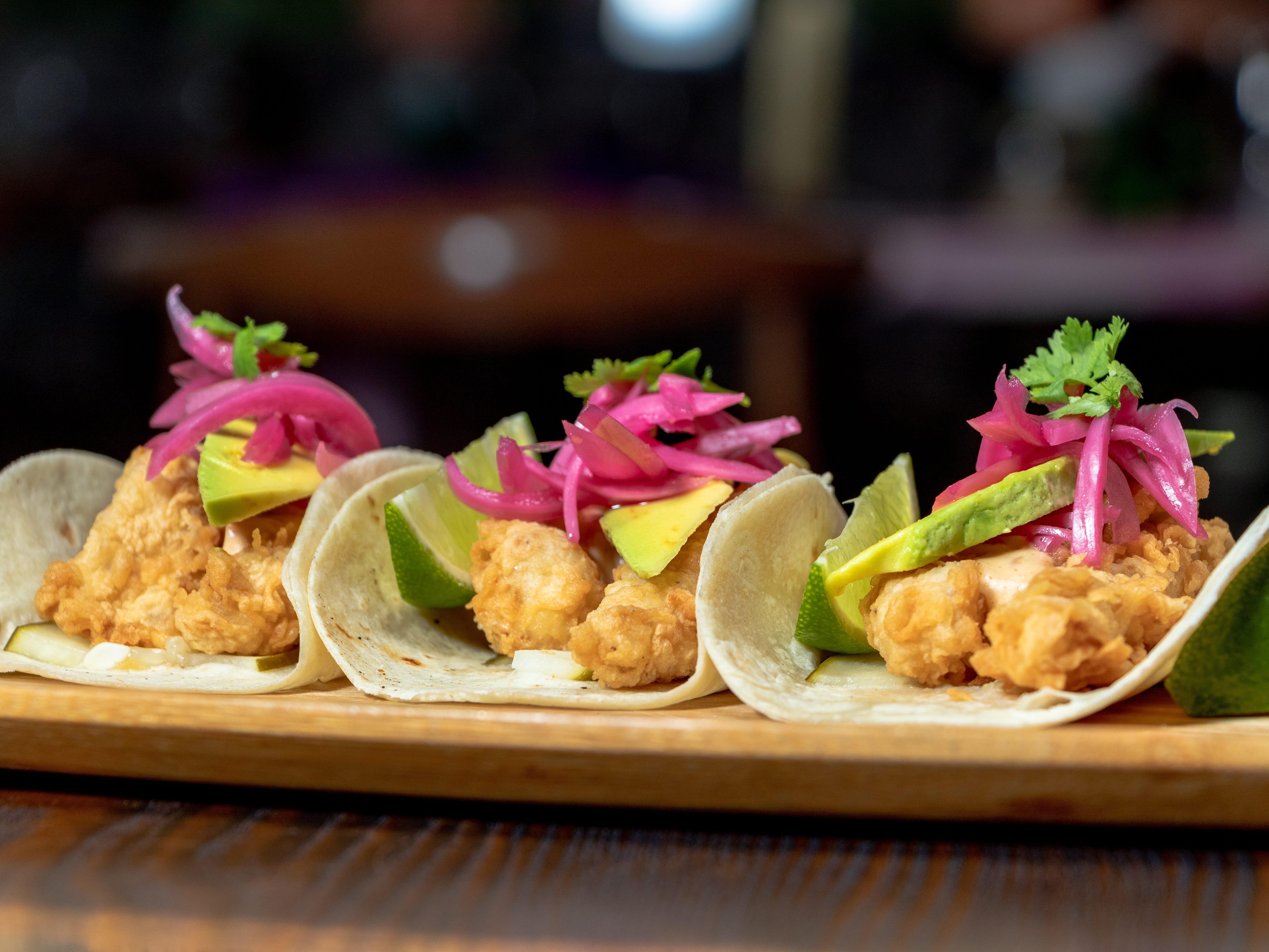 Three small tacos topped with pickled red onions, cilantro, avocado, and lime slices on a wooden tray.