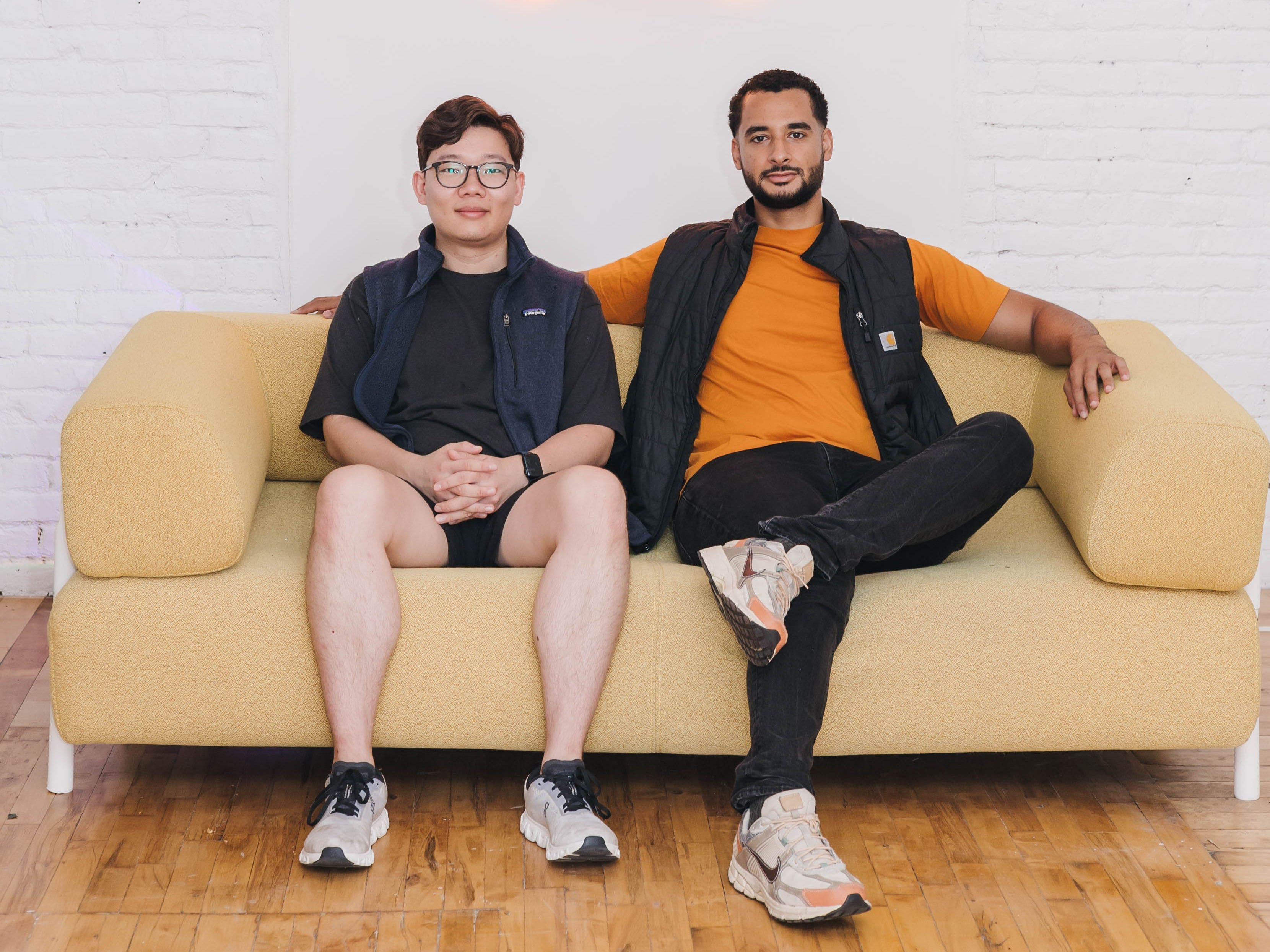 Zingage cofounders Daniel Tian and Victor Hunt, sitting on a yellow sofa in front of a white brick wall with the Zingage logo in neon lights.