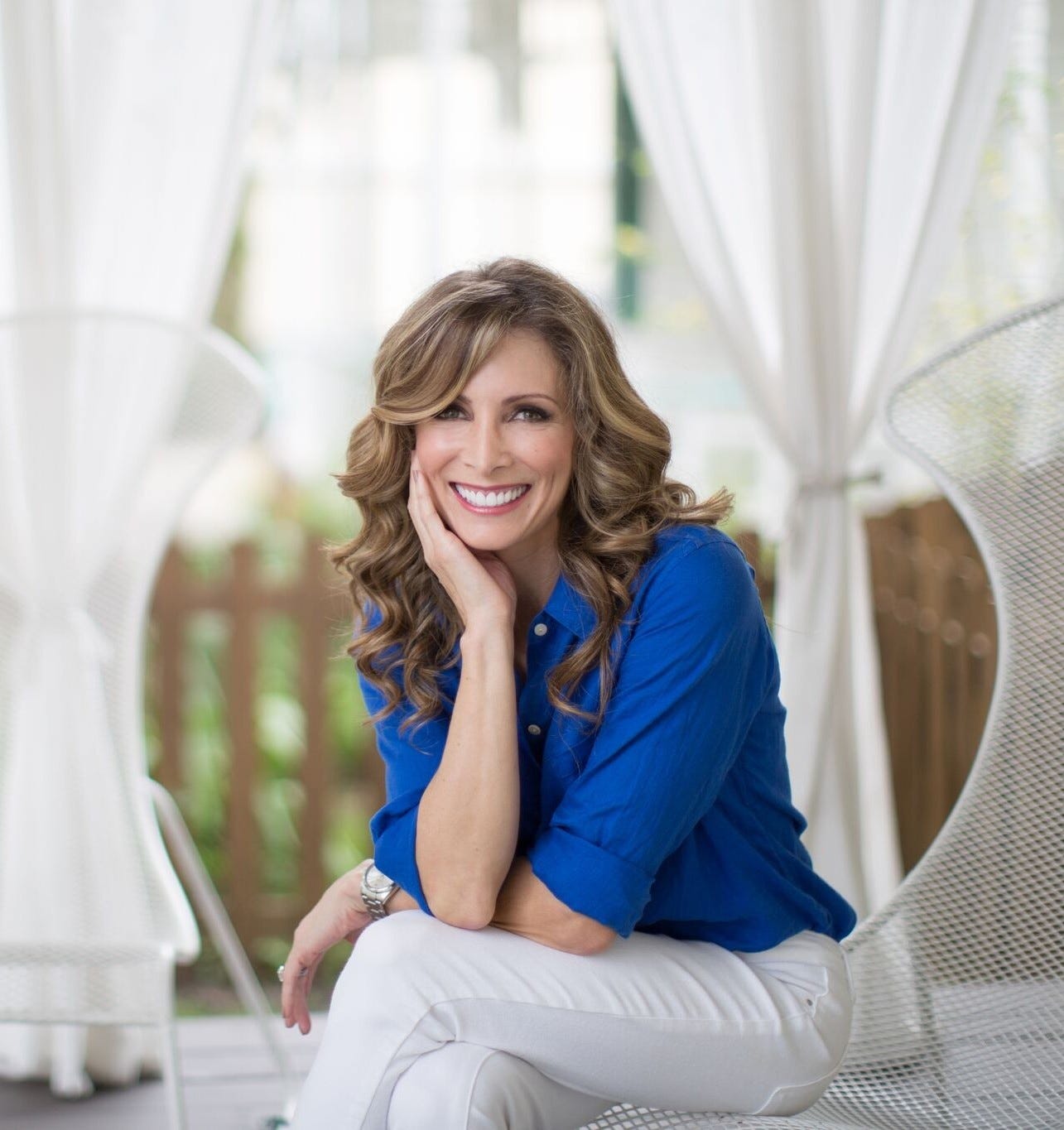 a portrait photo of Shannon Miller smiling at the camera in a blue blouse and white pants in a sunlit space