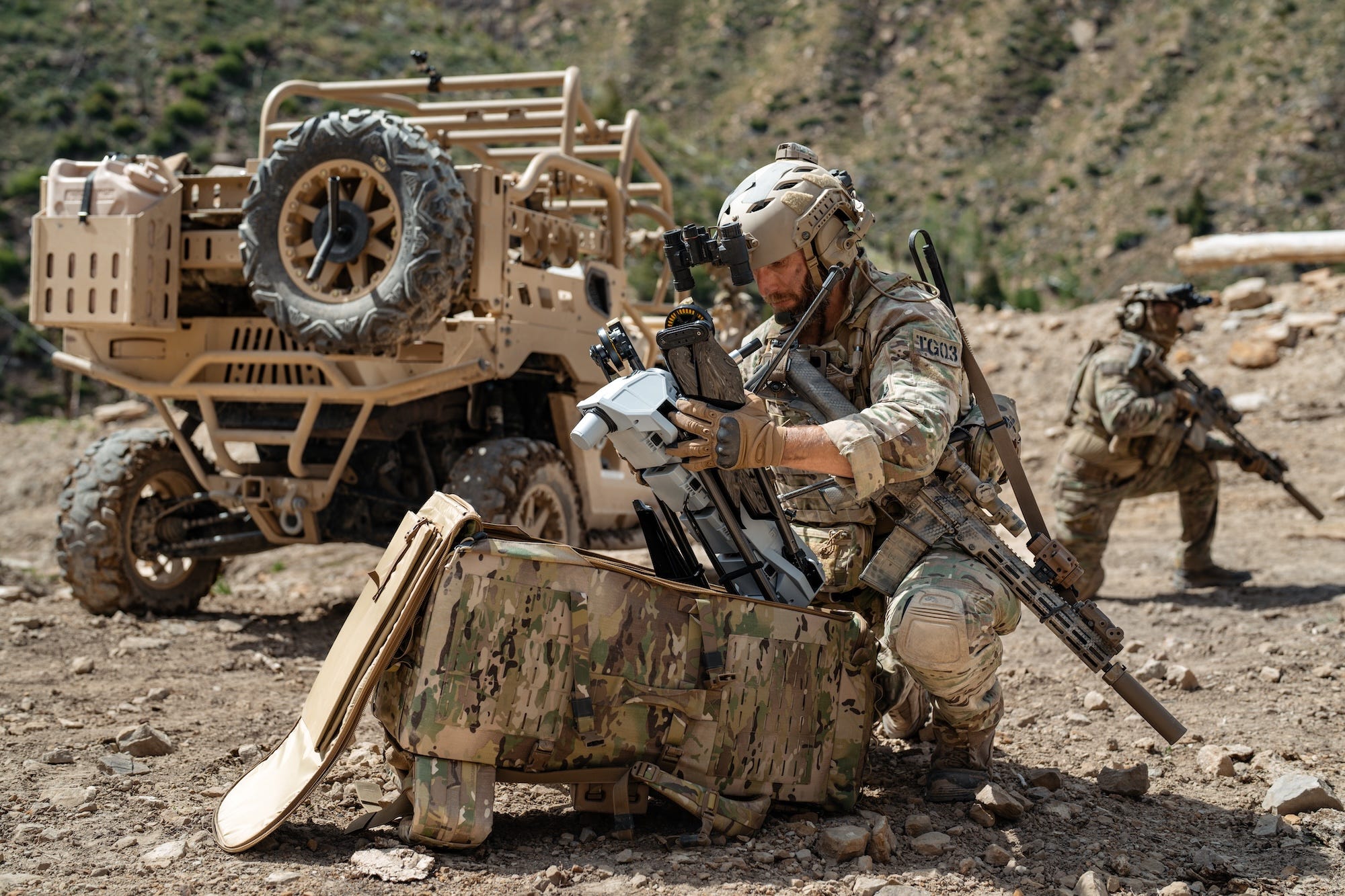 A man in camoflage gear squats and takes a pale-colored drone out of a camoflage backpack, with a vehicle and another person behind him, all outdoors
