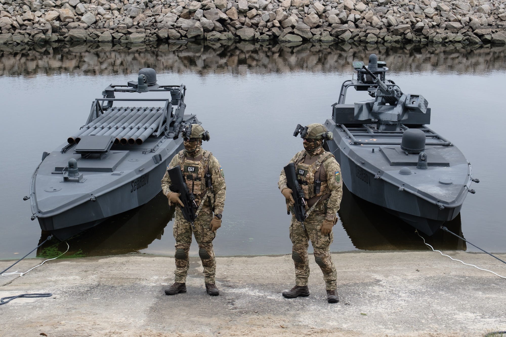 Two figures in camoflage gears stand on land beside two large boat-shaped grey drones in the water