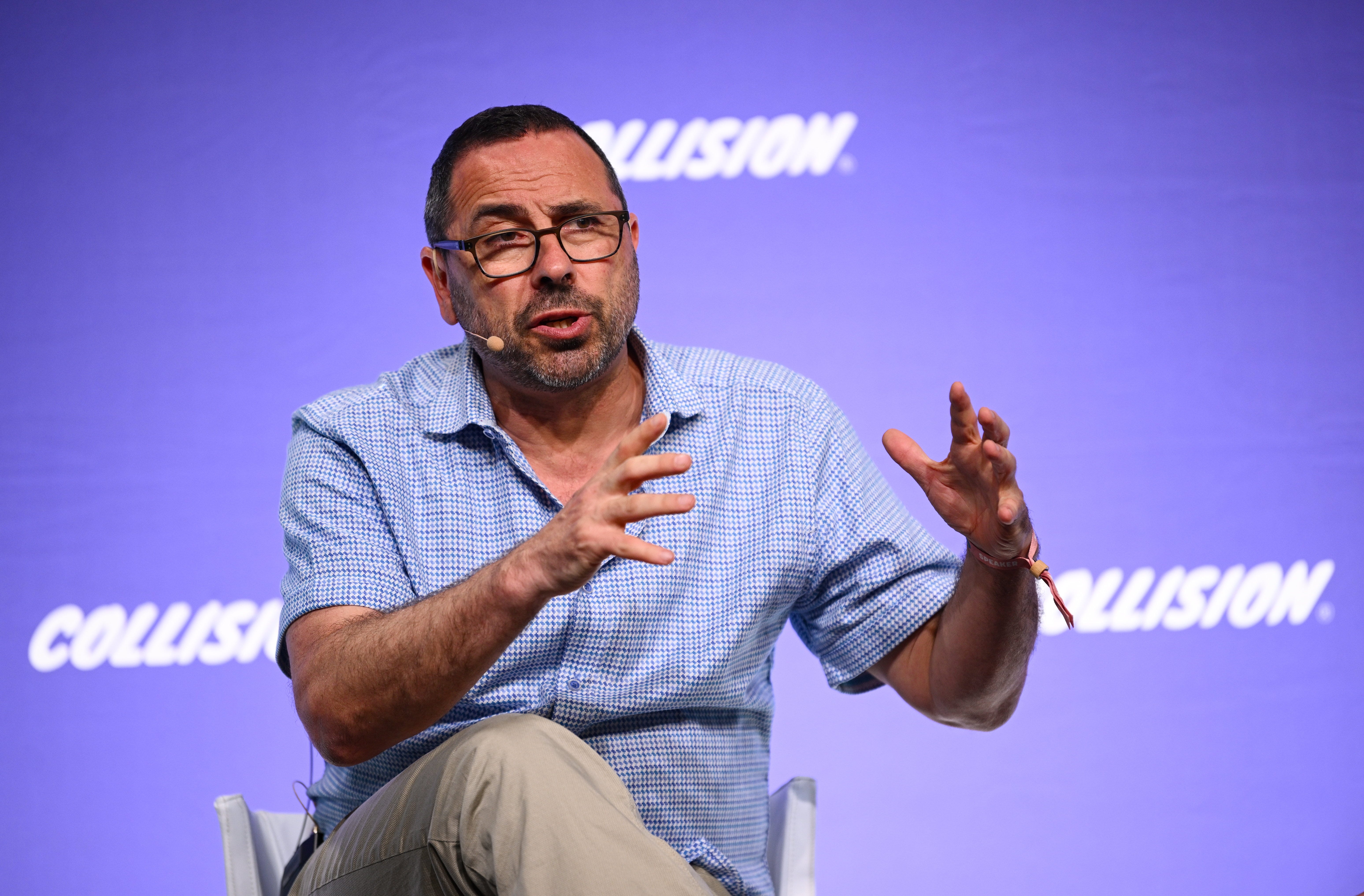 Andrew Feldman sits on a stage in casual clothes speaking and gesticulating wearing a head mic in front of a purple background that say