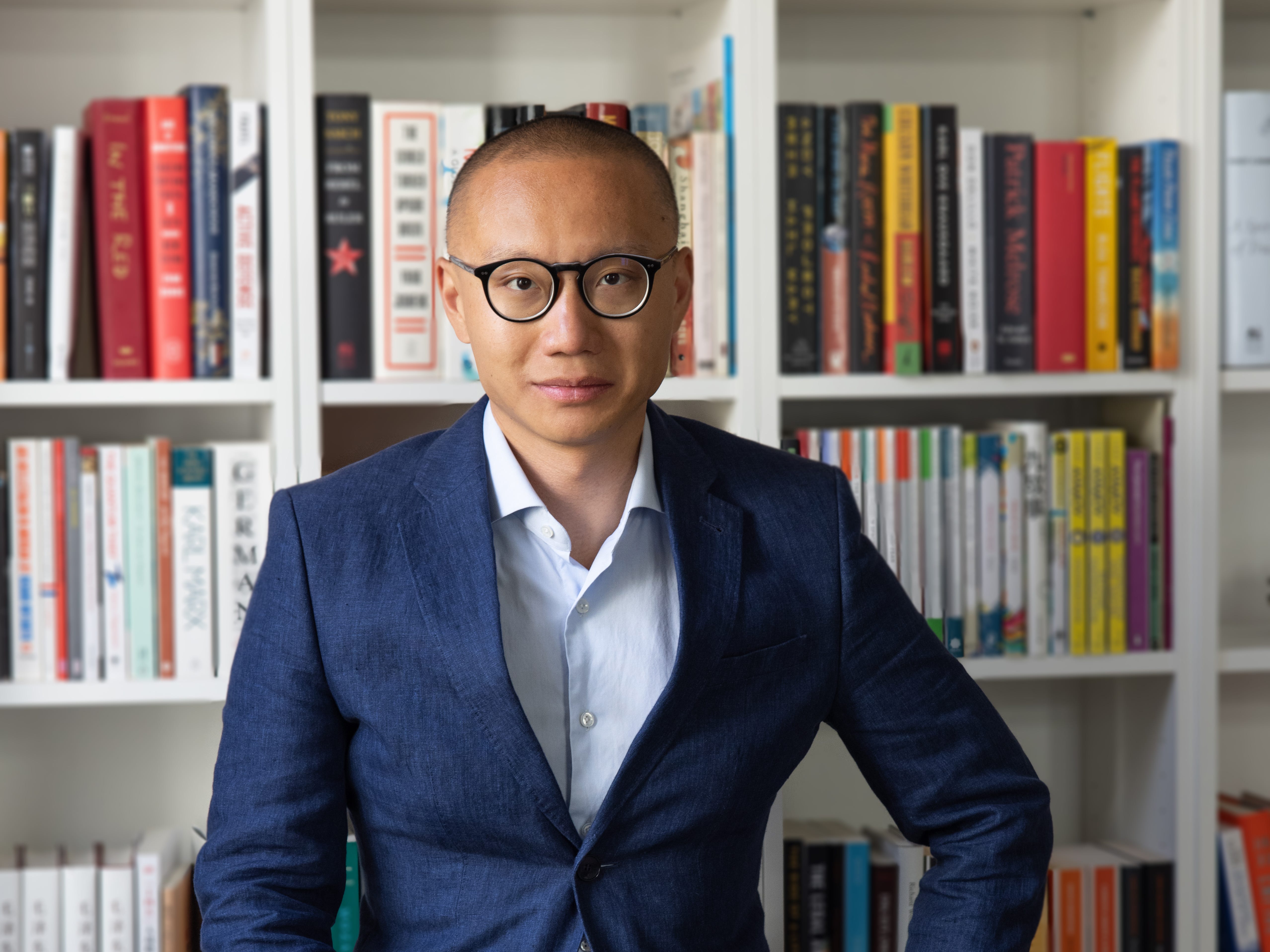 Dan Wang is wearing a blue jacket and sitting in front of his shelves.