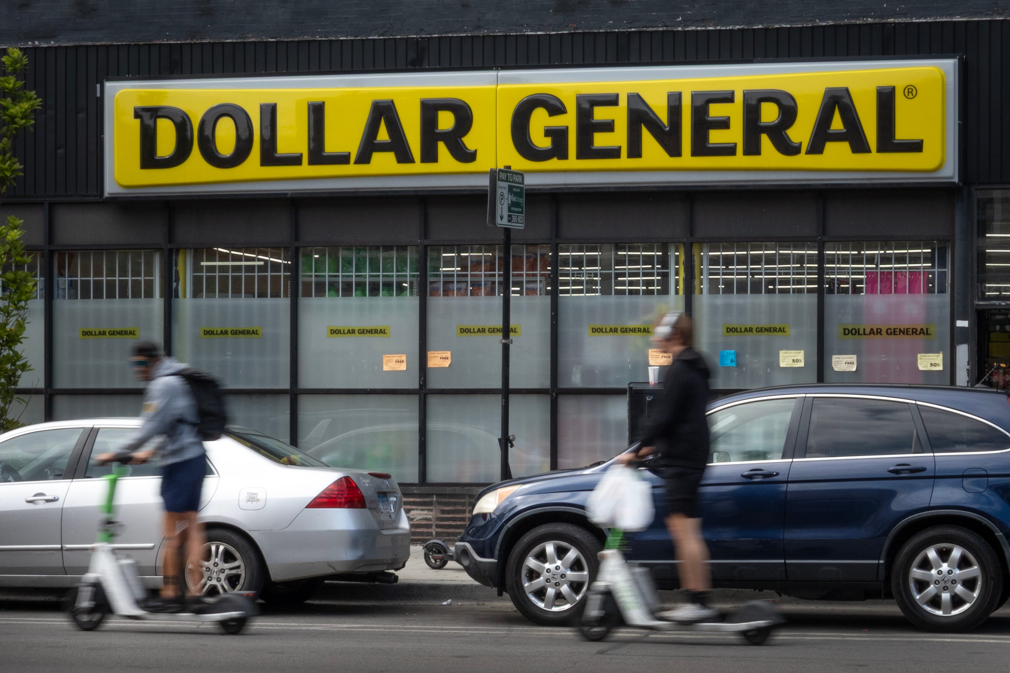 Two men ride by a Dollar General store on electric scooters while holding plastic bags