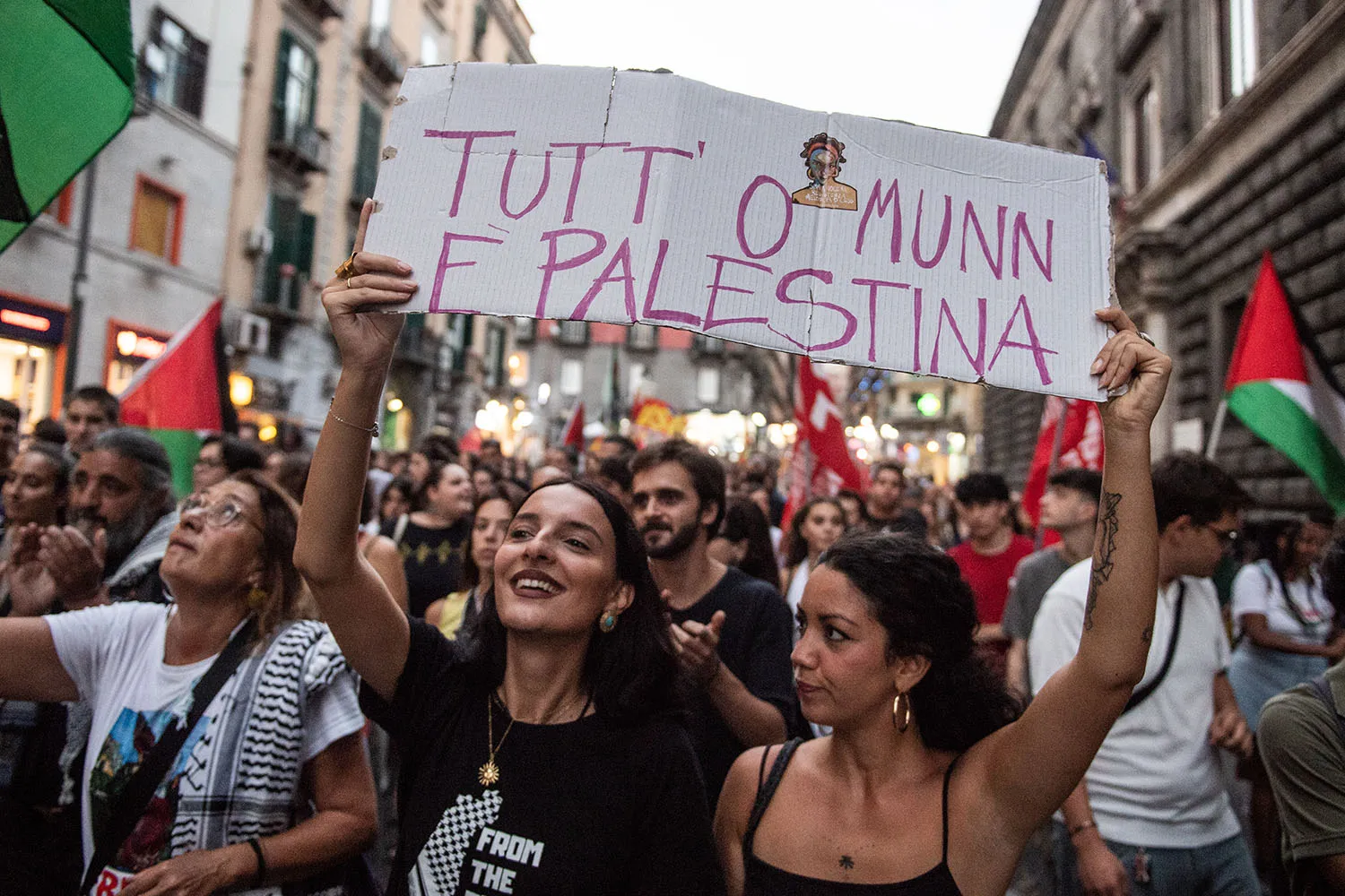 Two people hold up a cardboard sign. Behind them is a large crowd holding Palestinian flags.