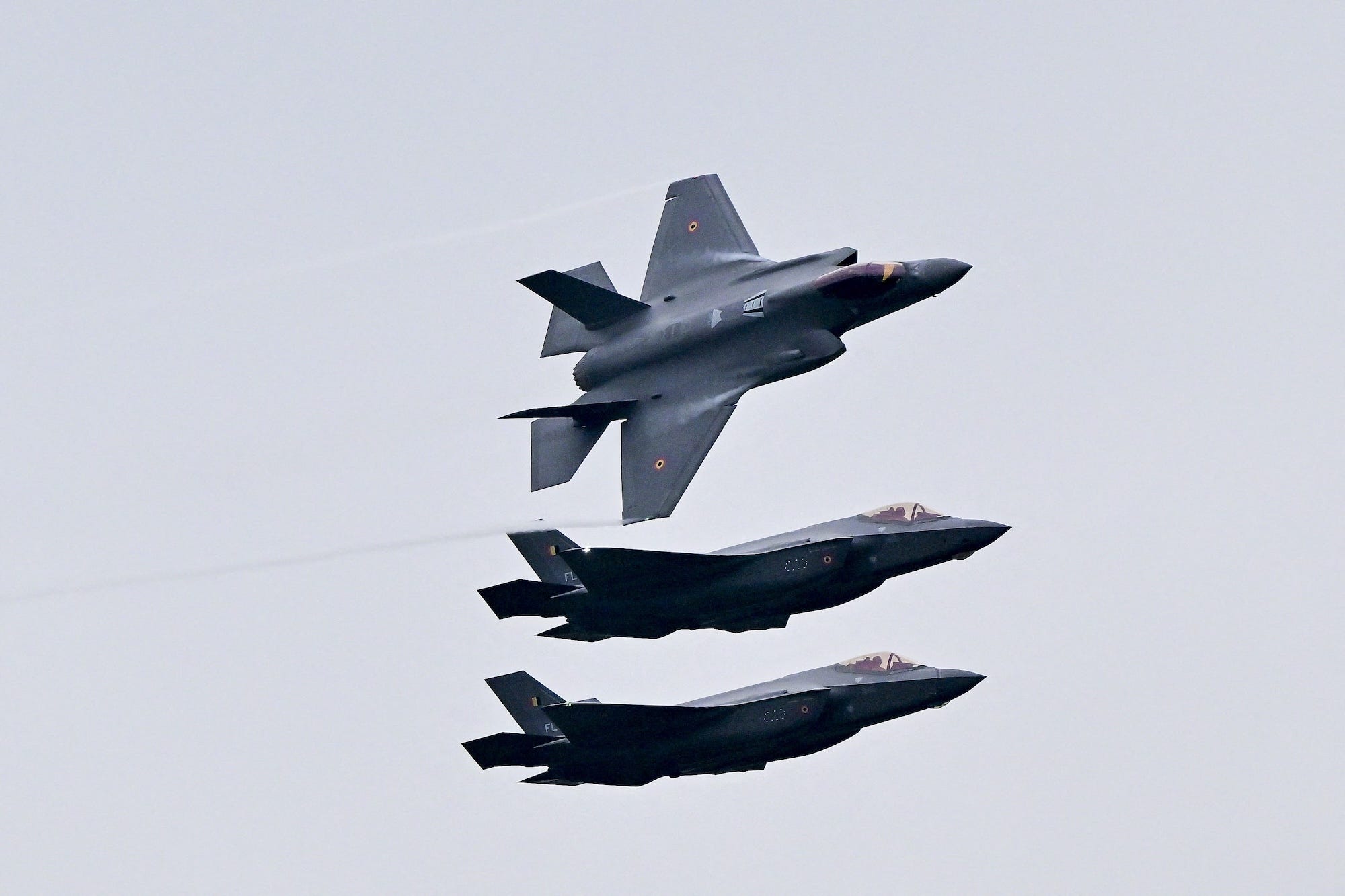 Three grey fighter jets in a grey sky, with the one on top turning on its side