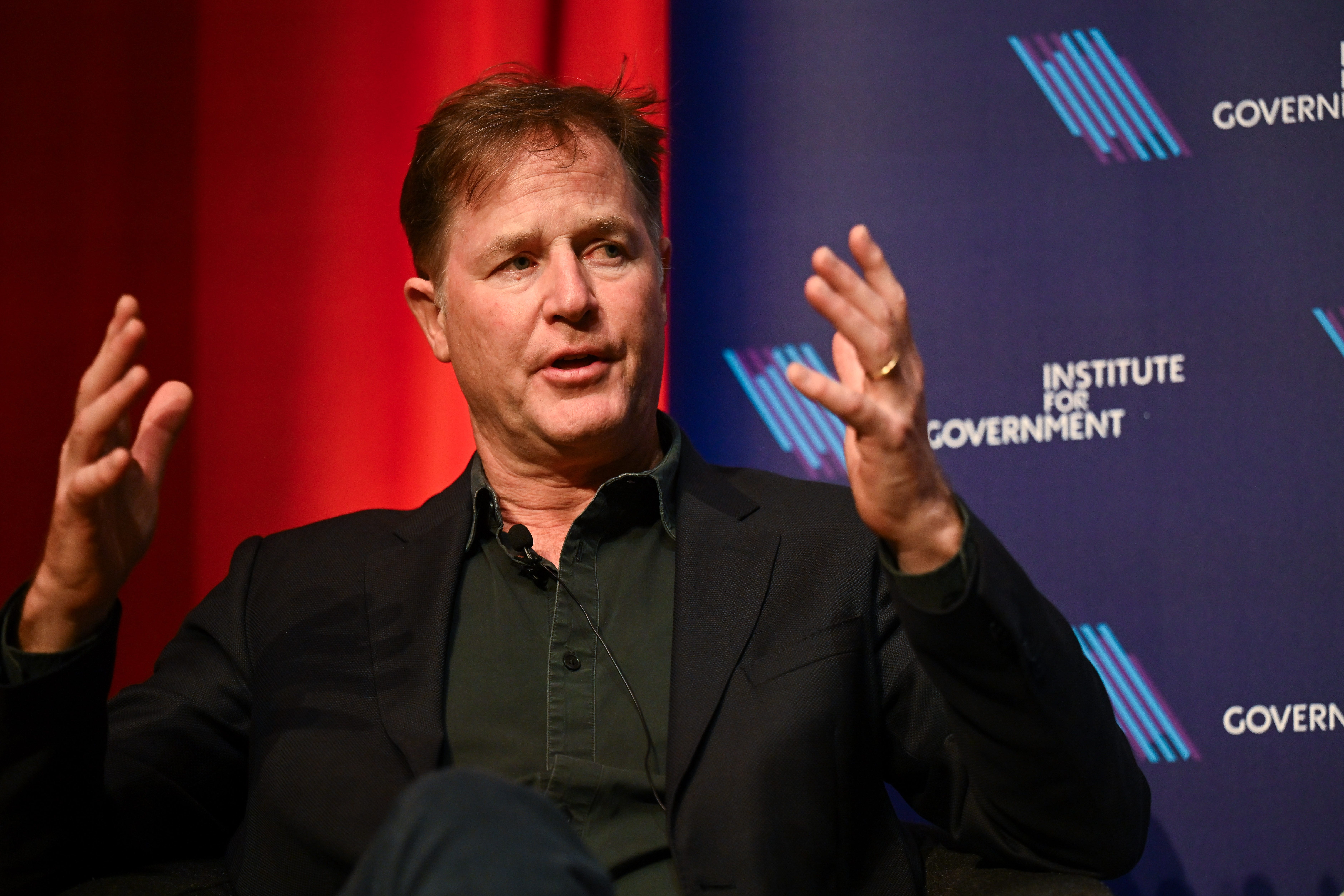 Nick Clegg is wearing a black shirt and a black blazer while gesturing with his hands.