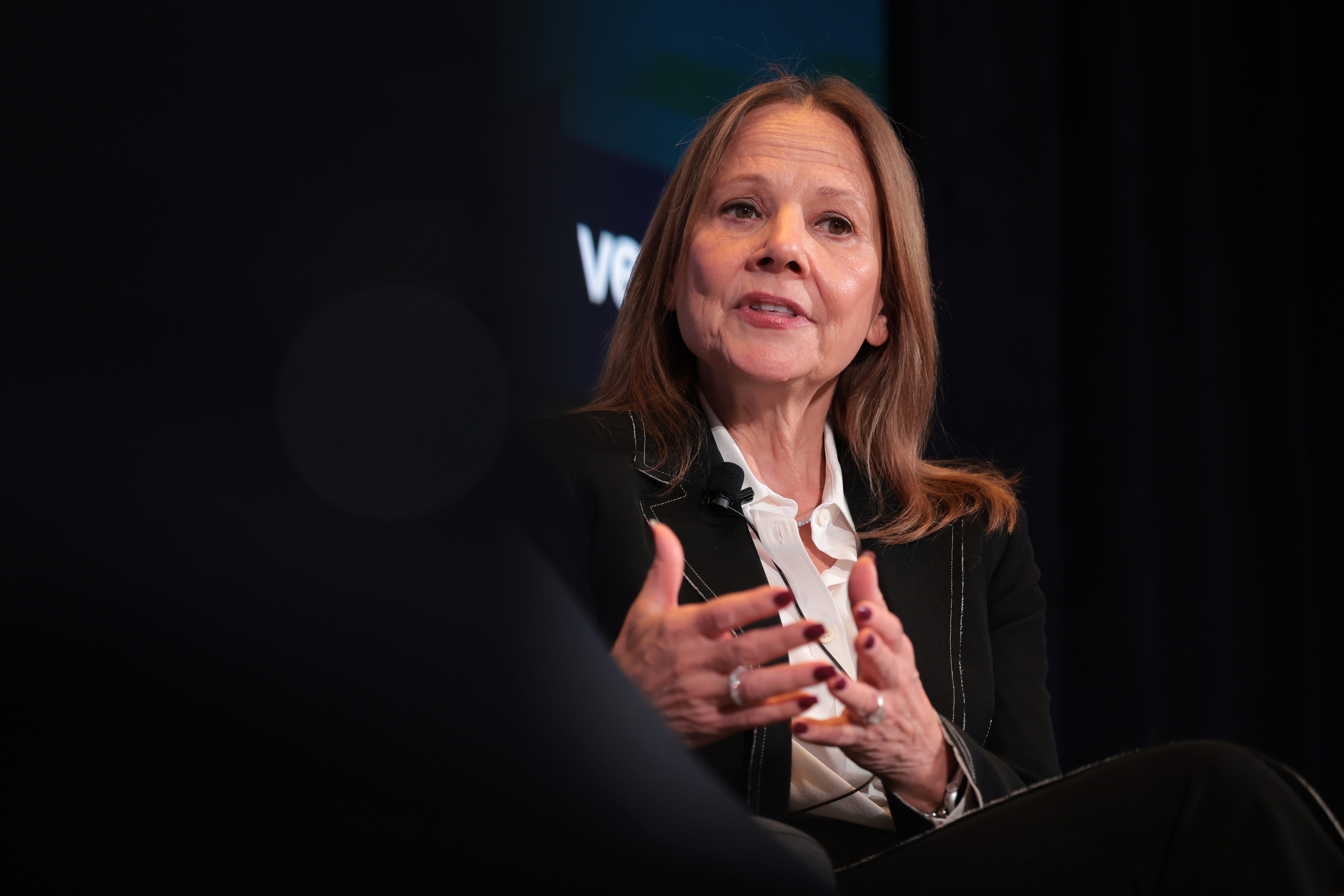 GM CEO Mary Barra is wearing a black jacket and gesturing with her two hands.